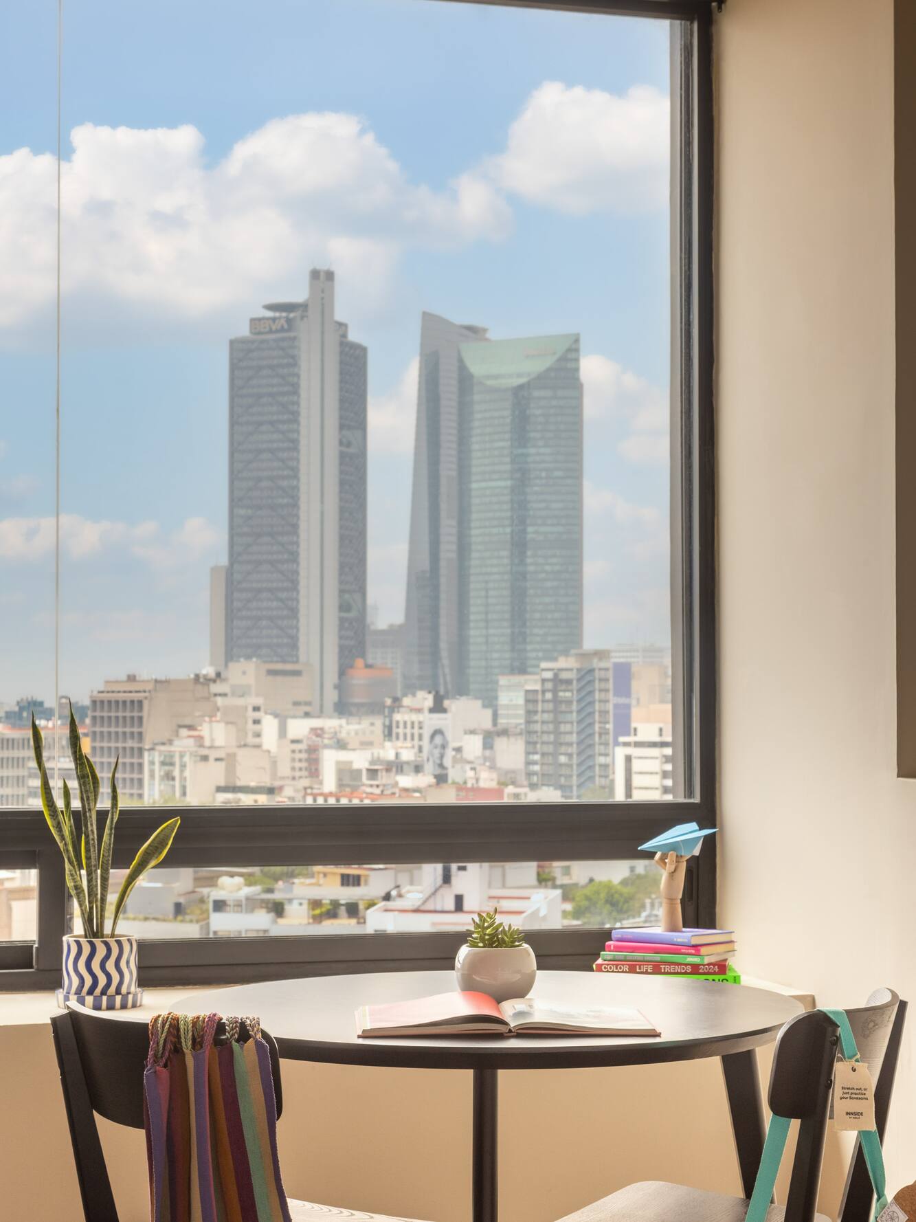 Mexico City apartment window view: modern table, plants, books, urban skyline.