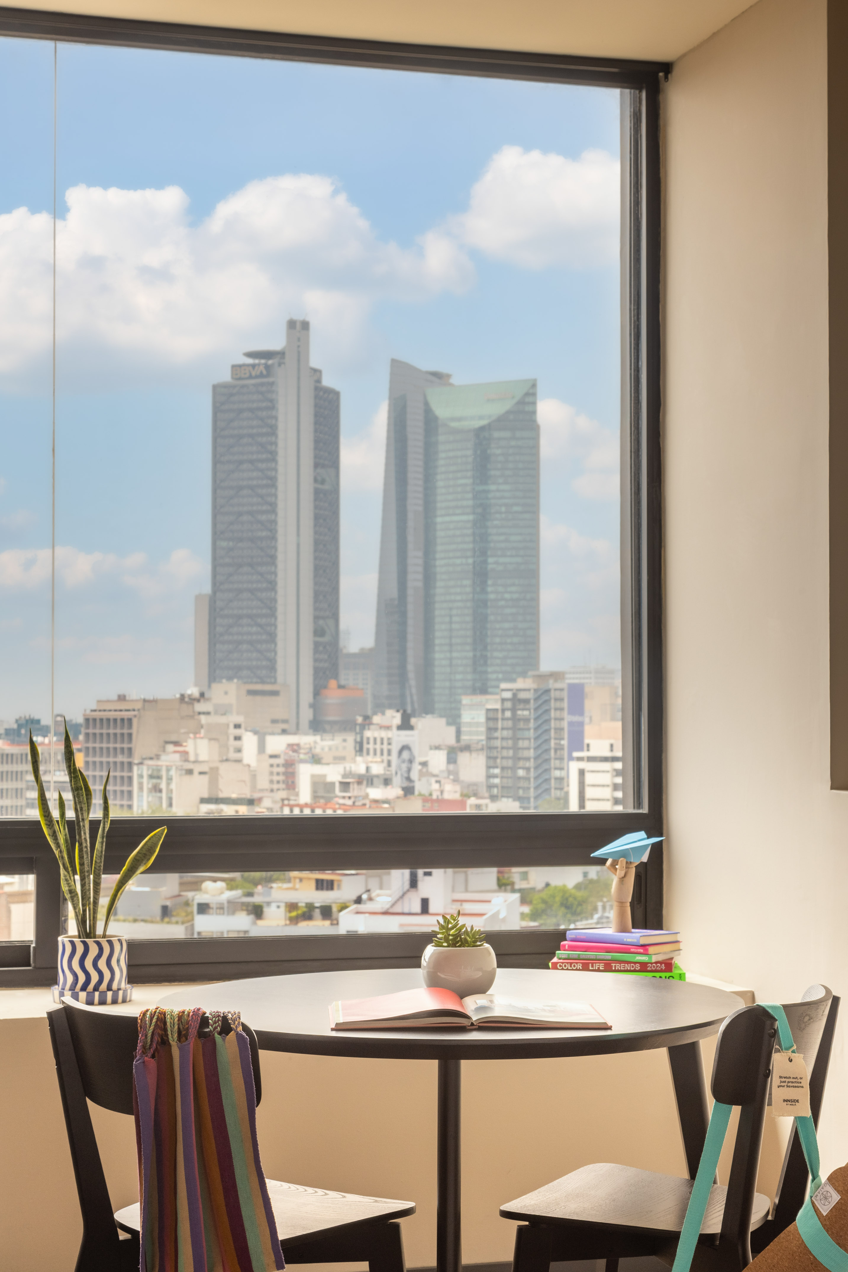 Mexico City apartment window view: modern table, plants, books, urban skyline.