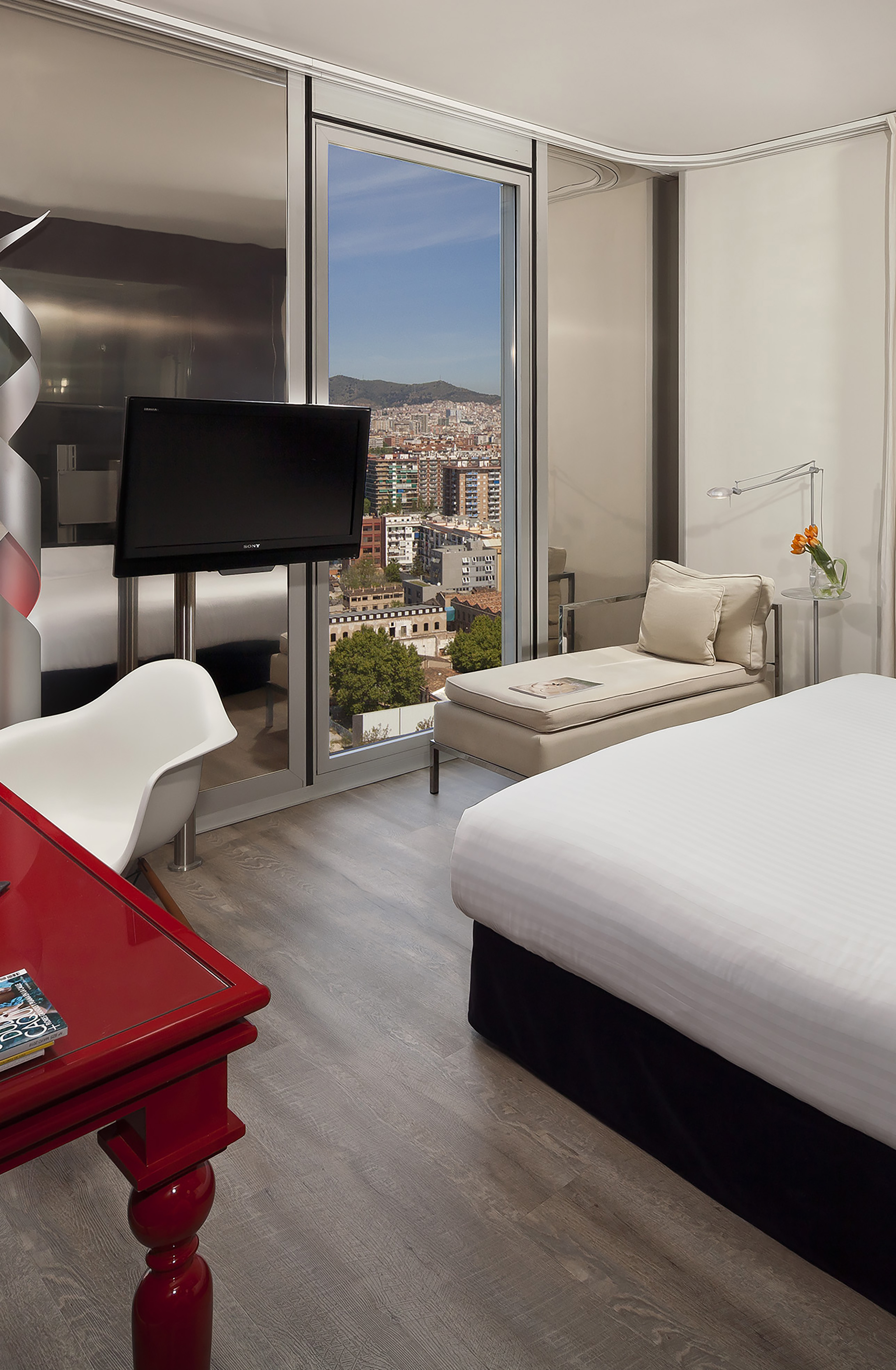 Modern Barcelona hotel room with city skyline view, bed, and chaise lounge.