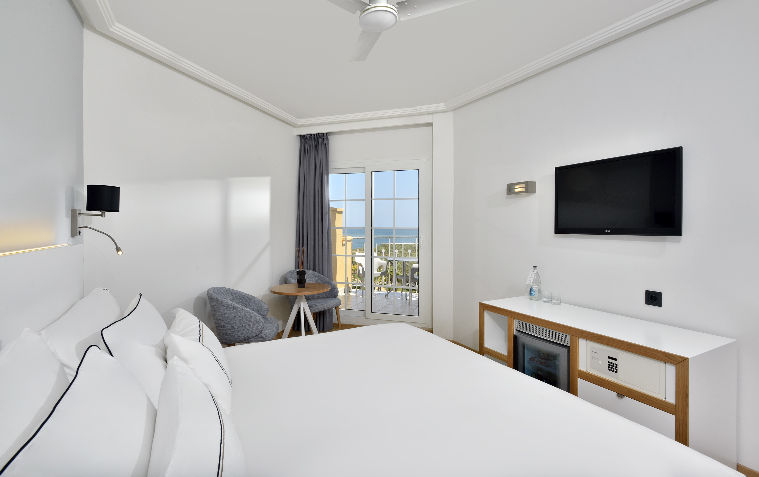 Modern hotel room with sea view balcony, white bed, and TV.