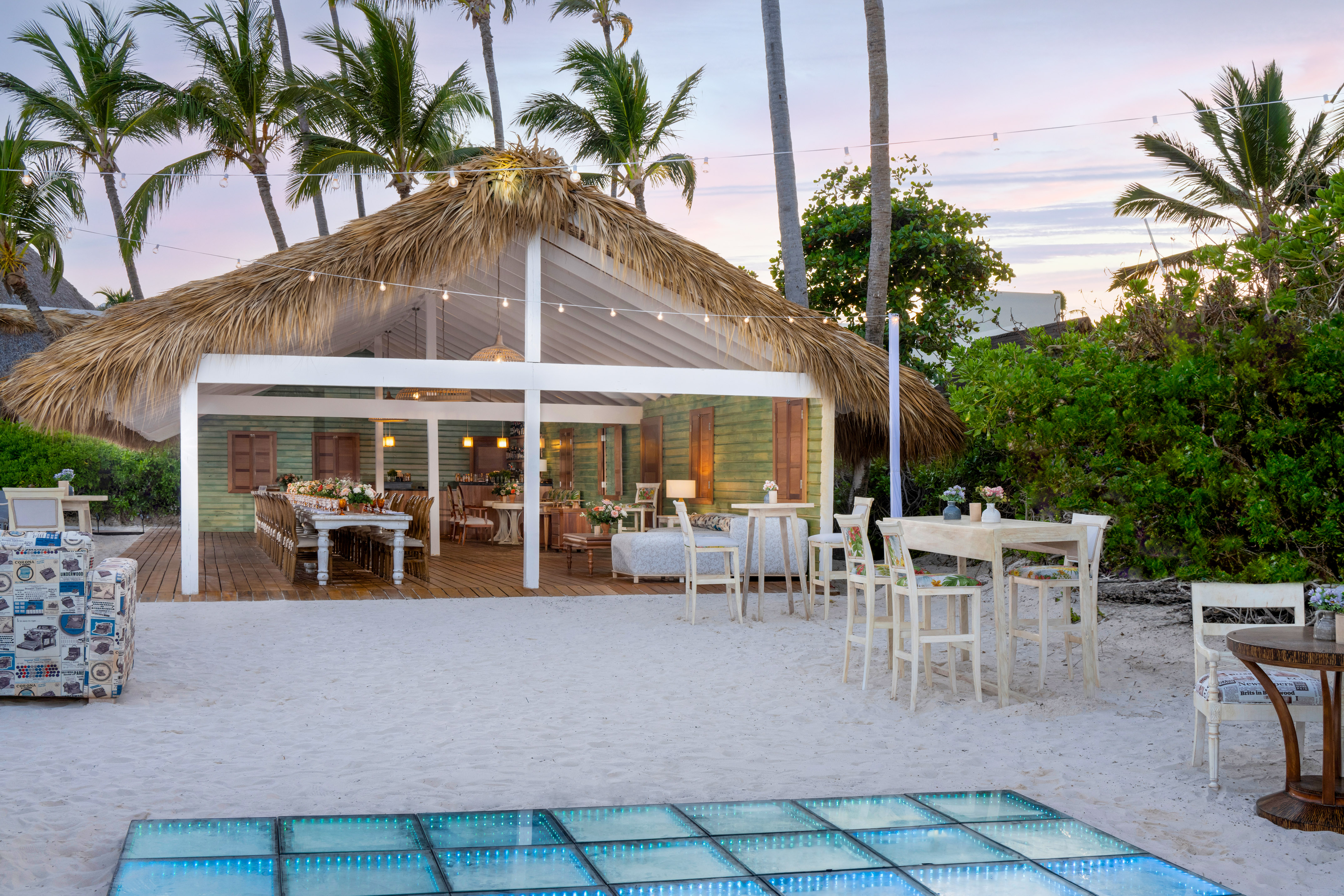 Tropical beach dining under thatch roof with palm trees and illuminated floor.
