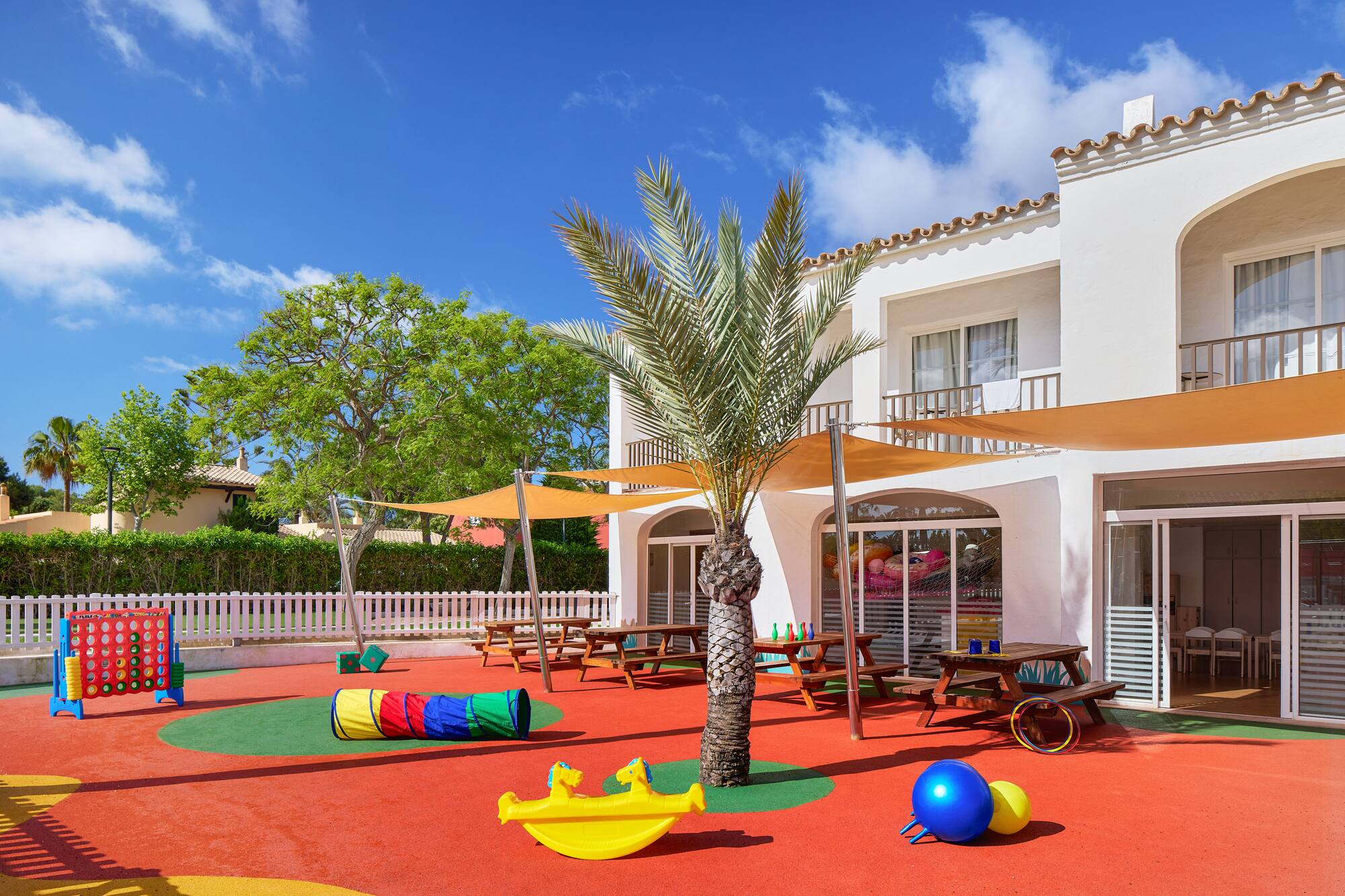 Outdoor kids playground with red floor, colorful equipment, hotel, and palm tree.