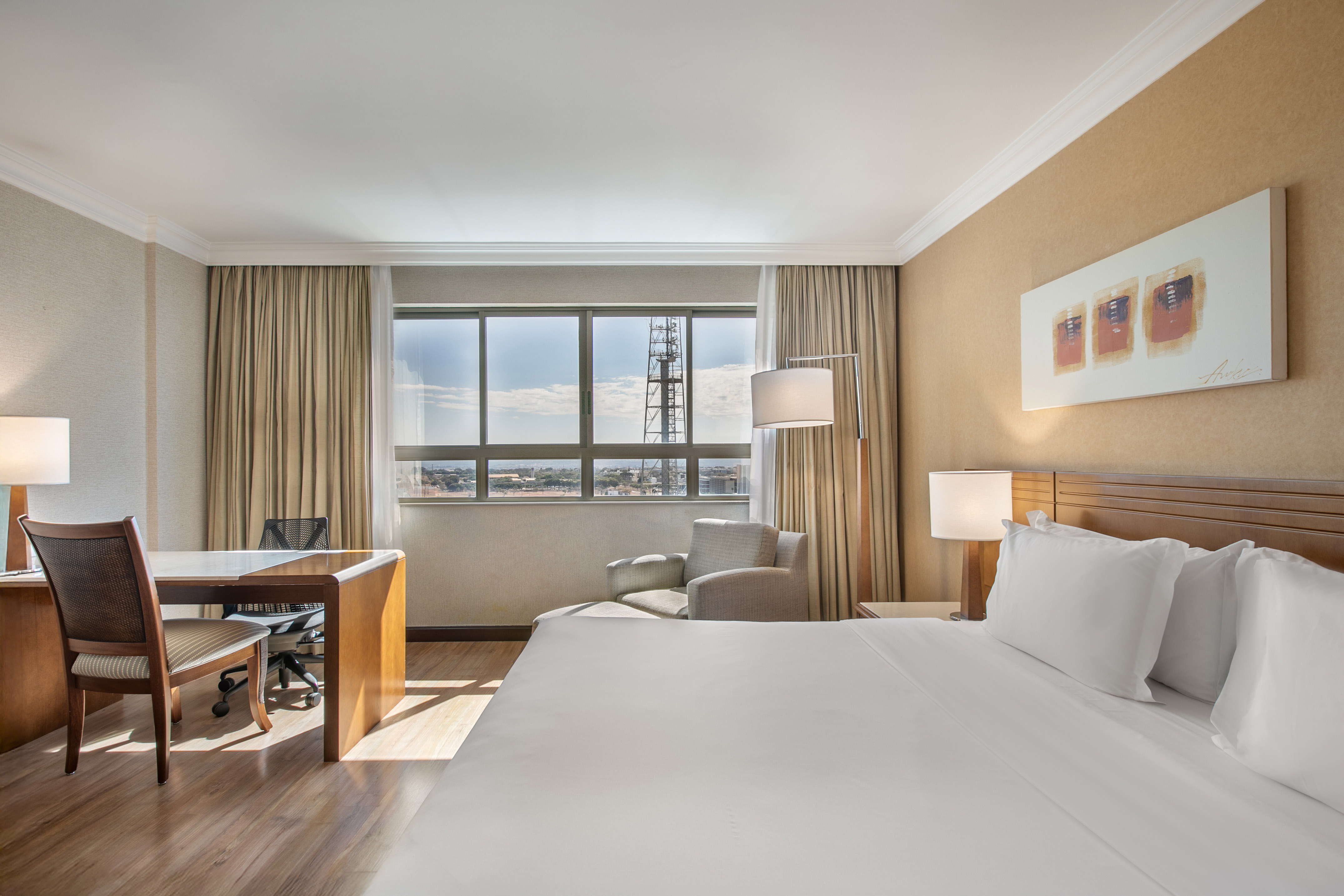 Hotel bedroom with large bed, desk, and city view from window.