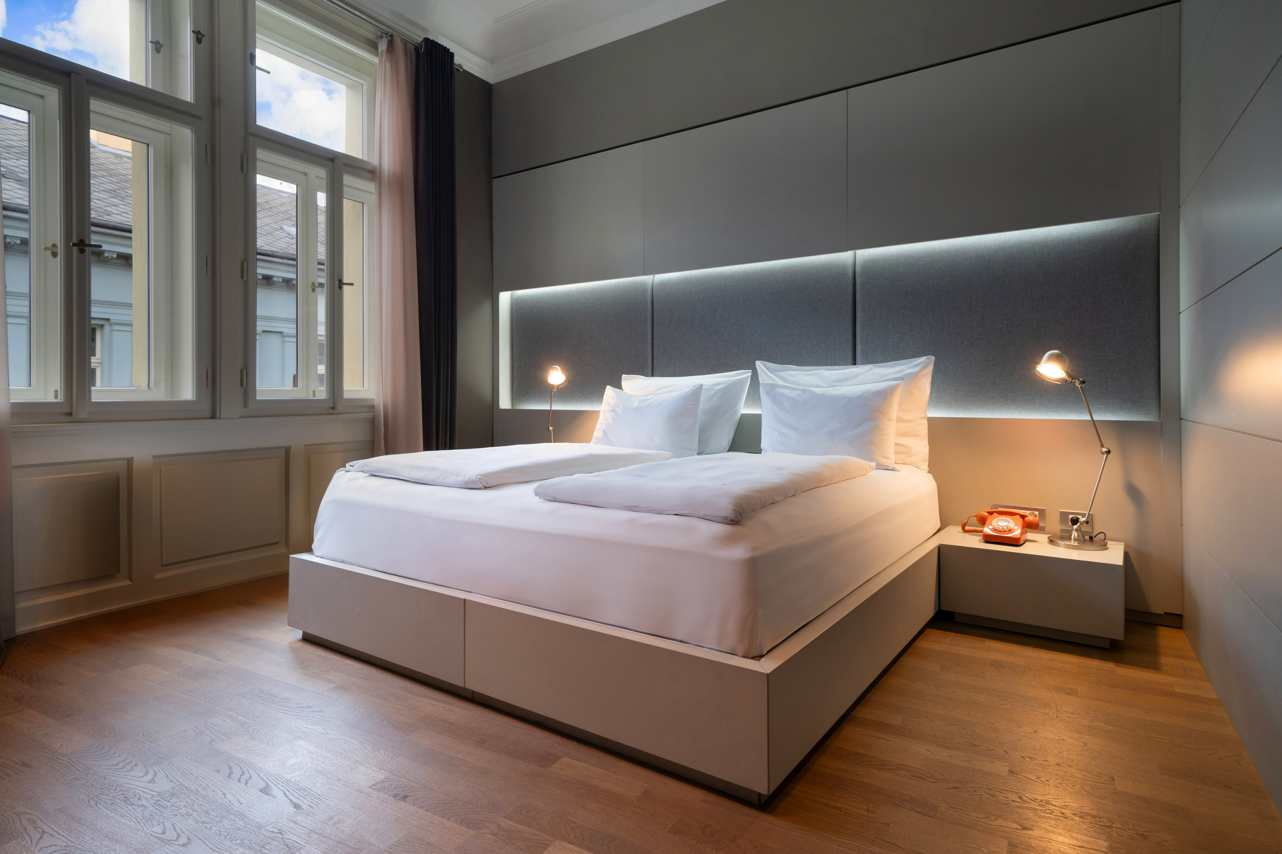 Modern hotel room with white bed, backlit headboard, wooden floor, large windows.