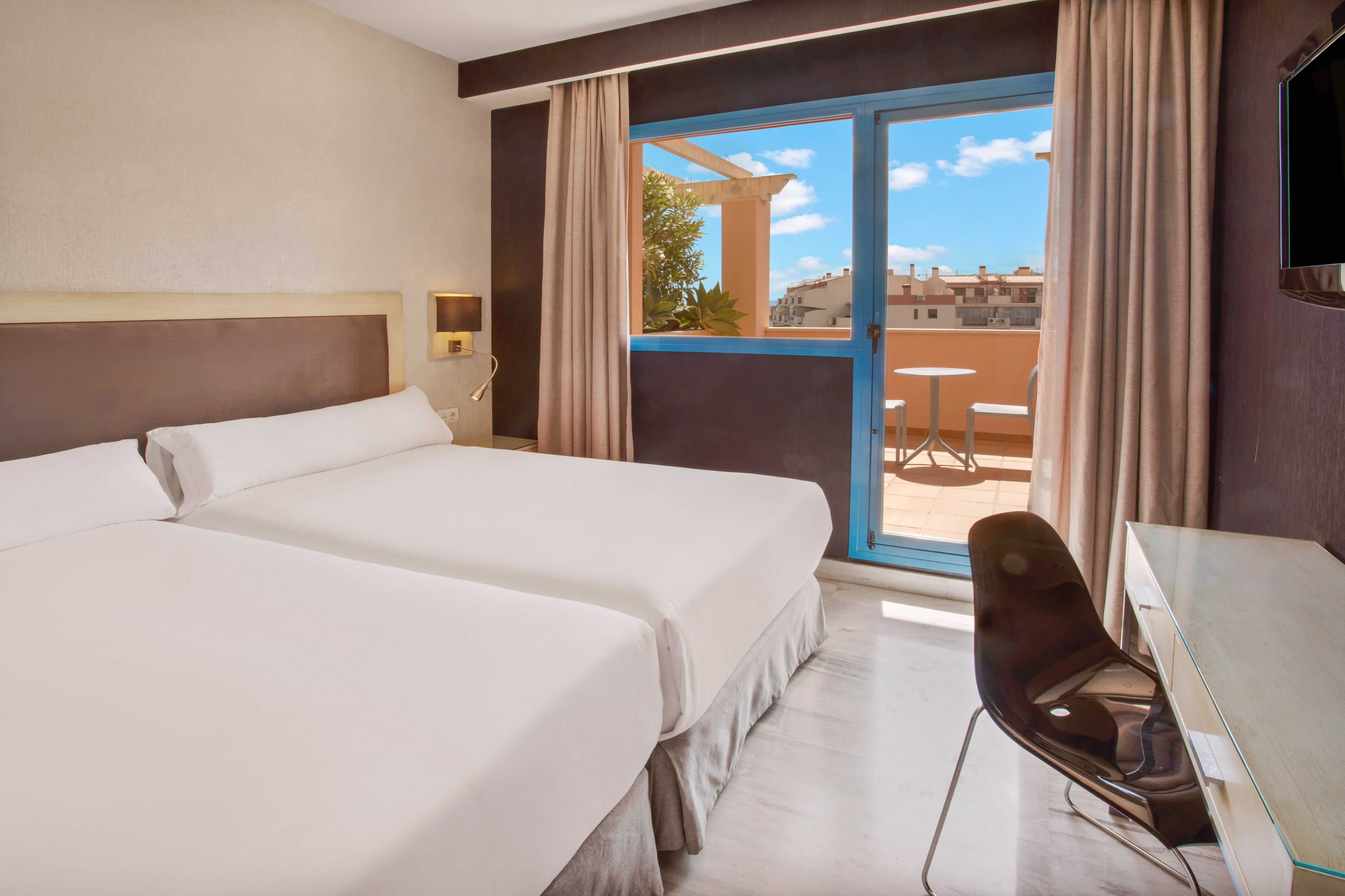 Bright hotel room with twin beds and balcony overlooking city rooftops.