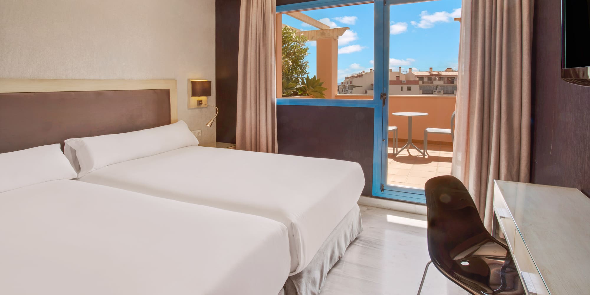Bright hotel room with twin beds and balcony overlooking city rooftops.