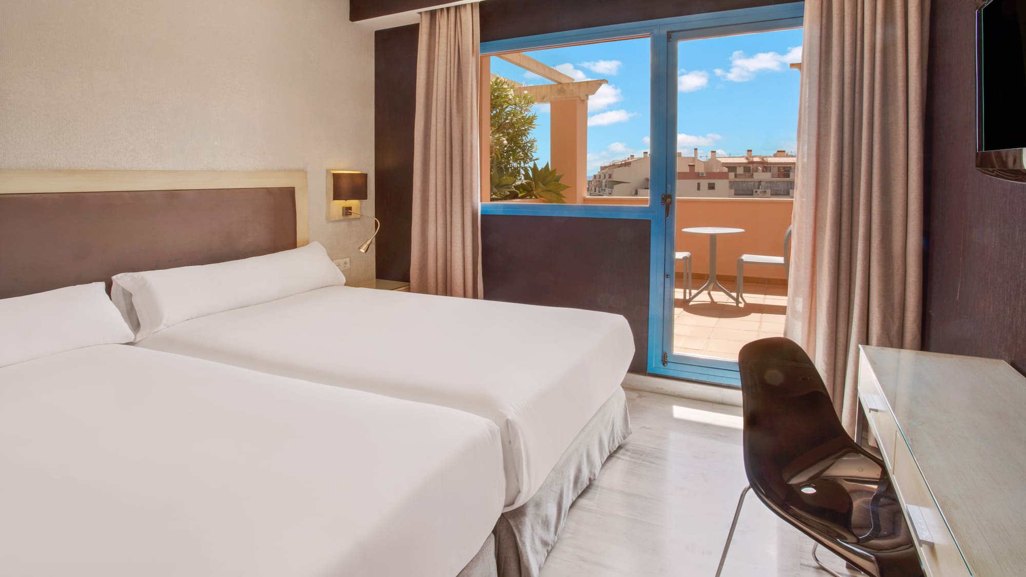 Bright hotel room with twin beds and balcony overlooking city rooftops.