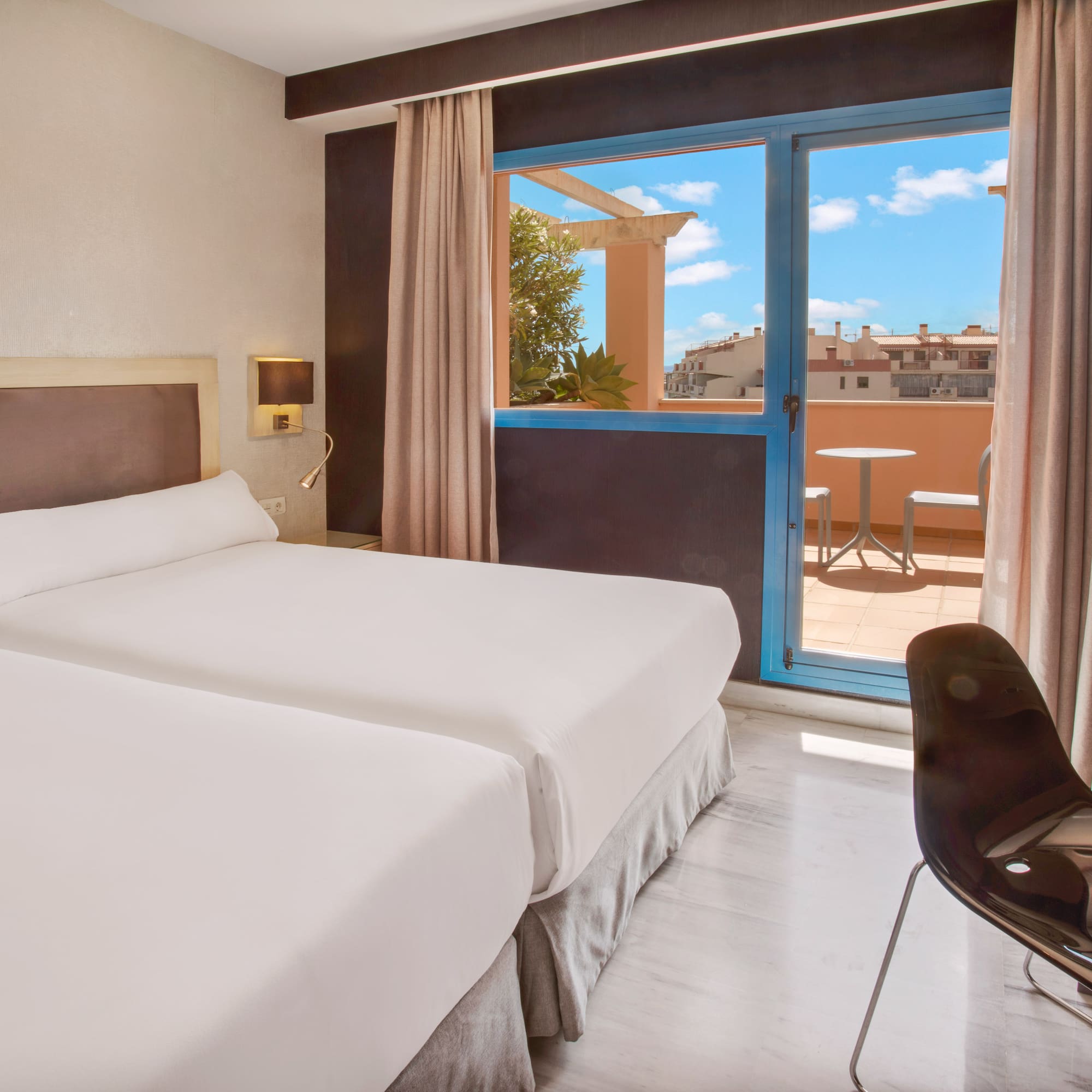 Bright hotel room with twin beds and balcony overlooking city rooftops.