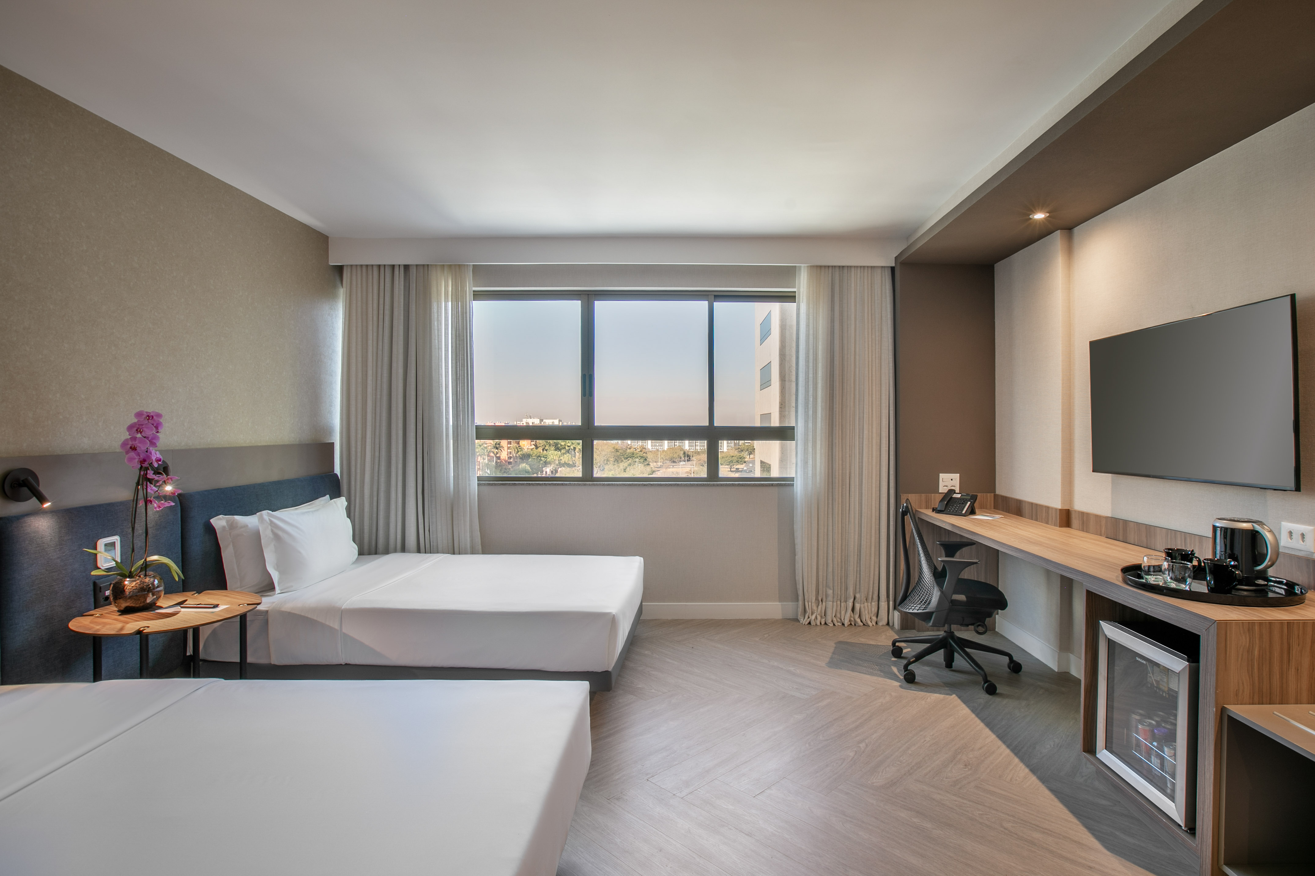 Modern hotel room with two twin beds, desk, TV, minibar, city view.