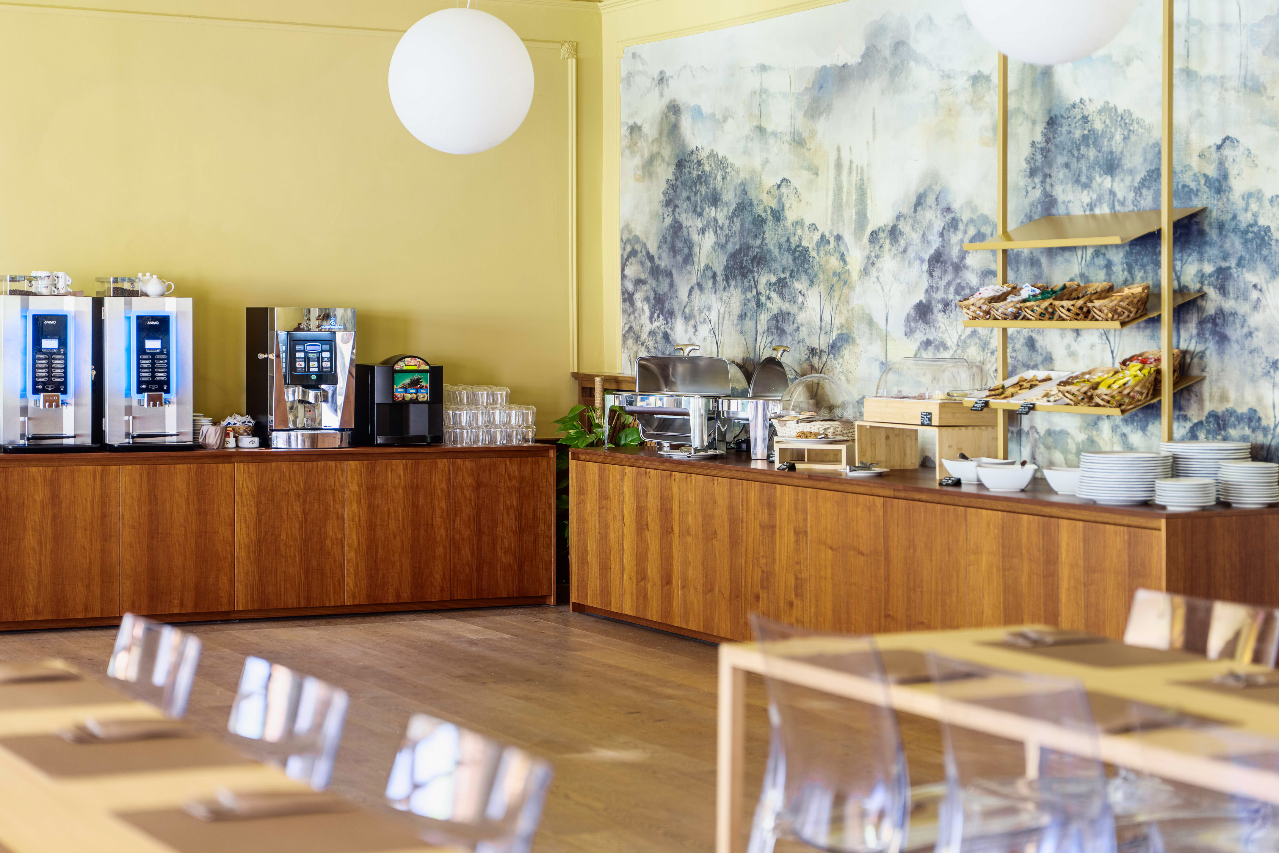 Hotel breakfast buffet with coffee, pastries, hot dishes, and scenic wall mural.