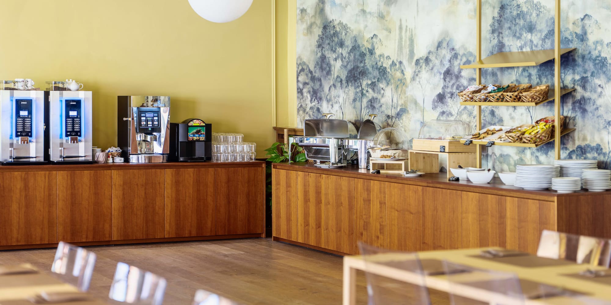 Hotel breakfast buffet with coffee, pastries, hot dishes, and scenic wall mural.