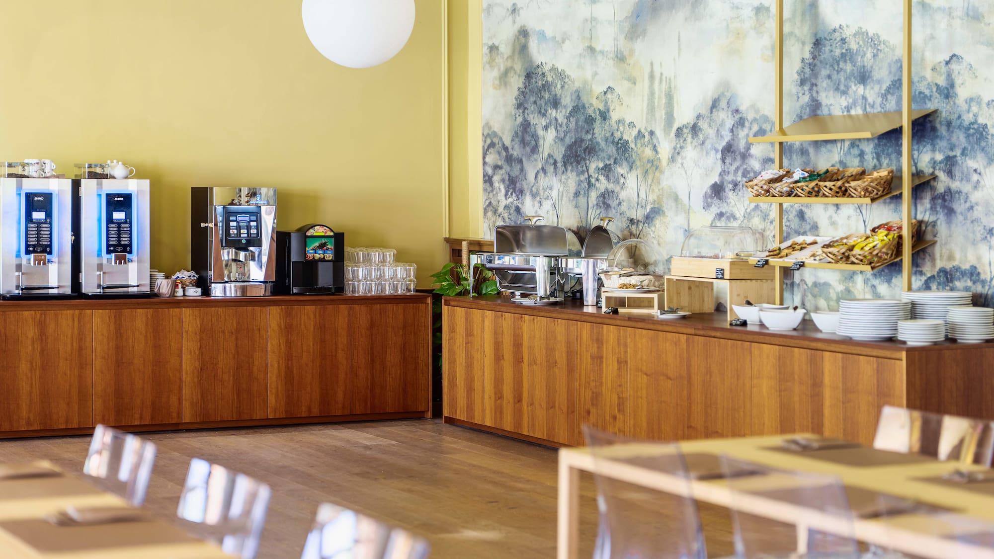 Hotel breakfast buffet with coffee, pastries, hot dishes, and scenic wall mural.