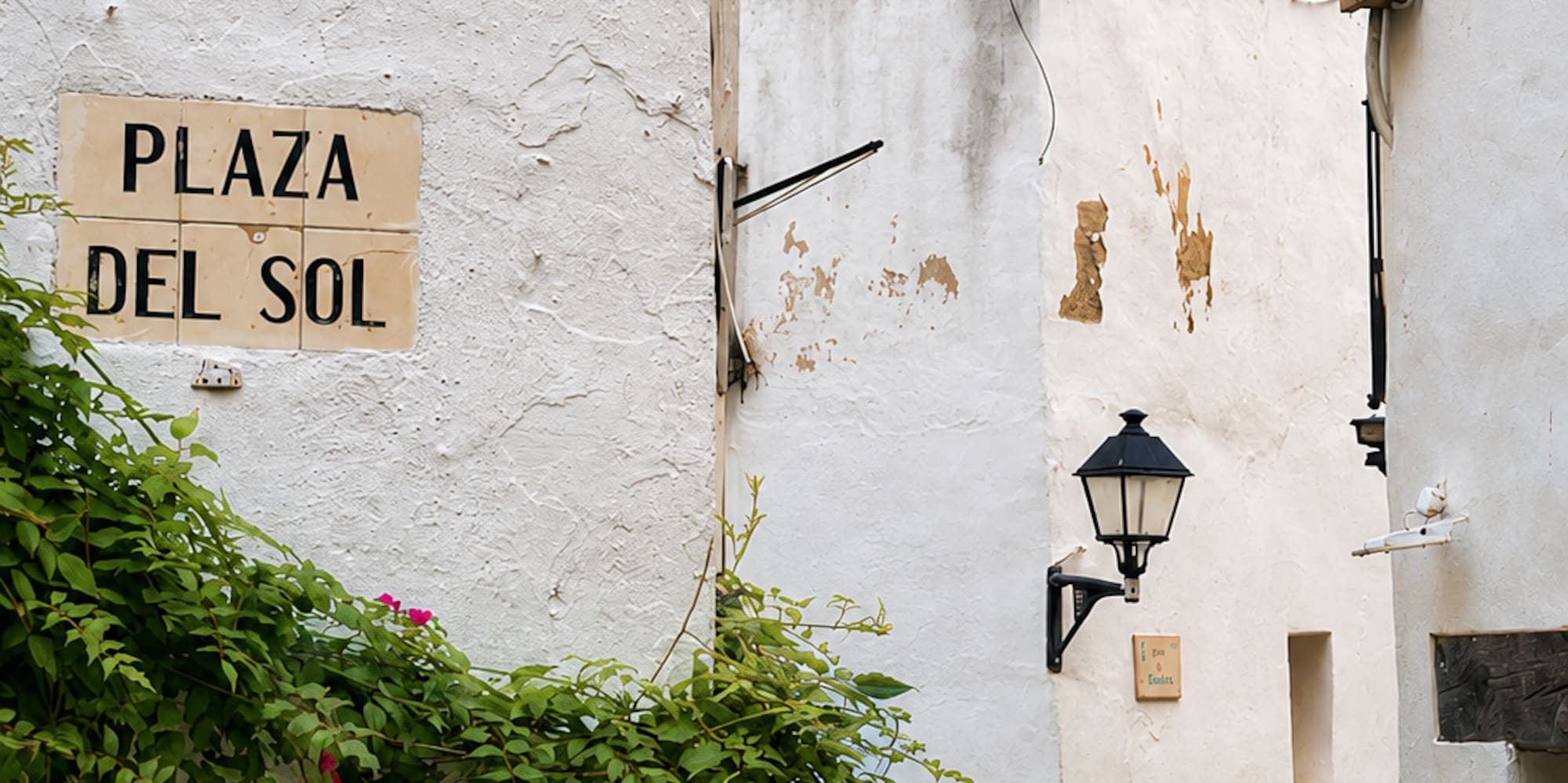White Mediterranean alley with 'Plaza del Sol' sign, green vines, street lamp.