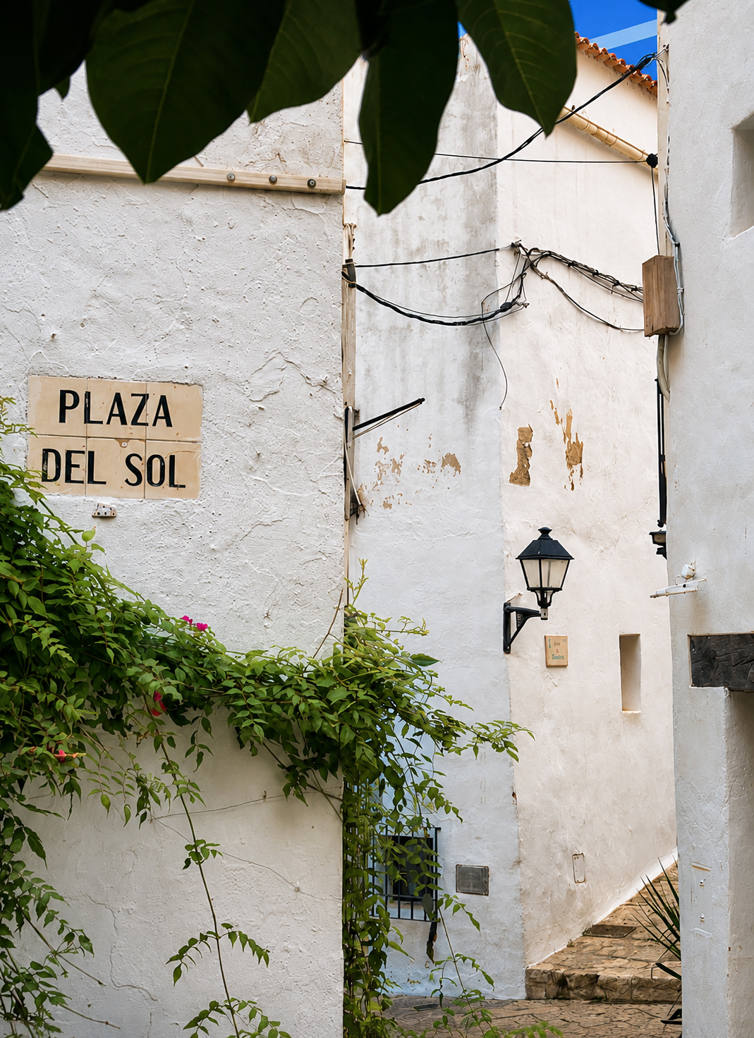 White Mediterranean alley with 'Plaza del Sol' sign, green vines, street lamp.
