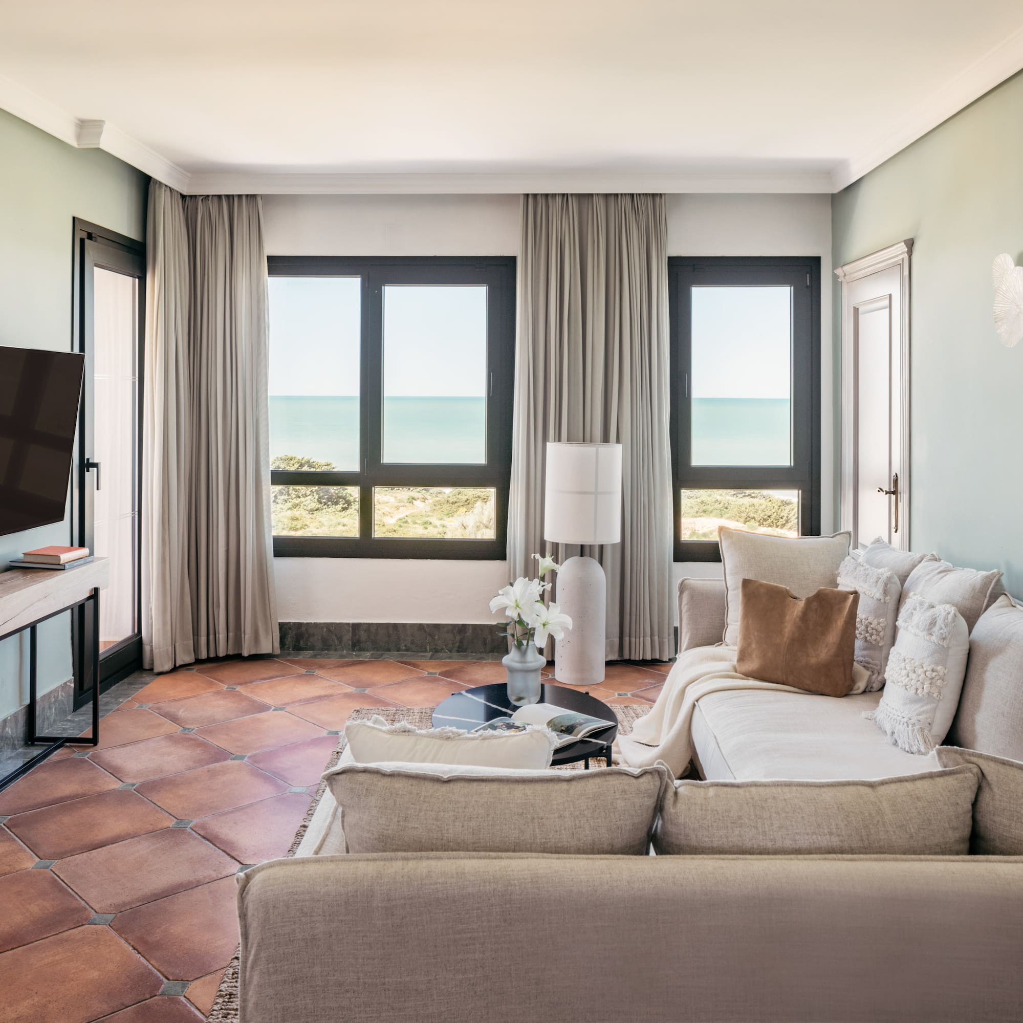 Coastal living room with sea view, light sofa, TV, and terracotta tiles.