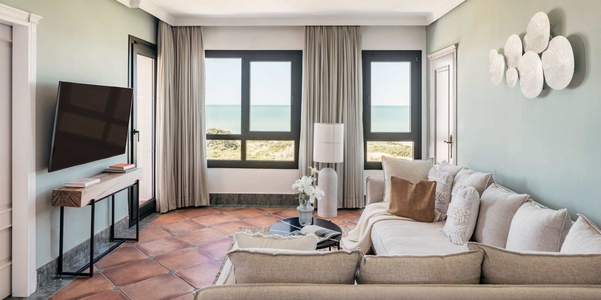 Coastal living room with sea view, light sofa, TV, and terracotta tiles.