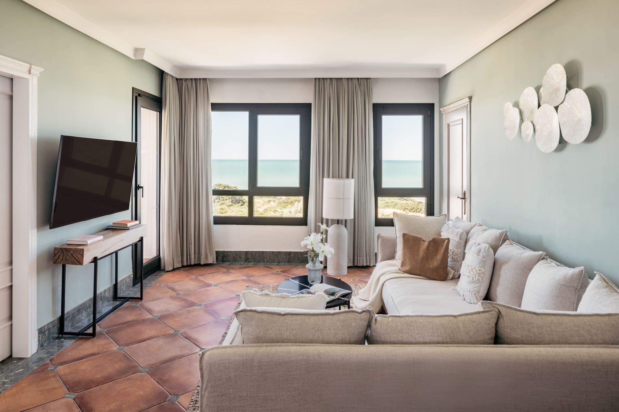 Coastal living room with sea view, light sofa, TV, and terracotta tiles.