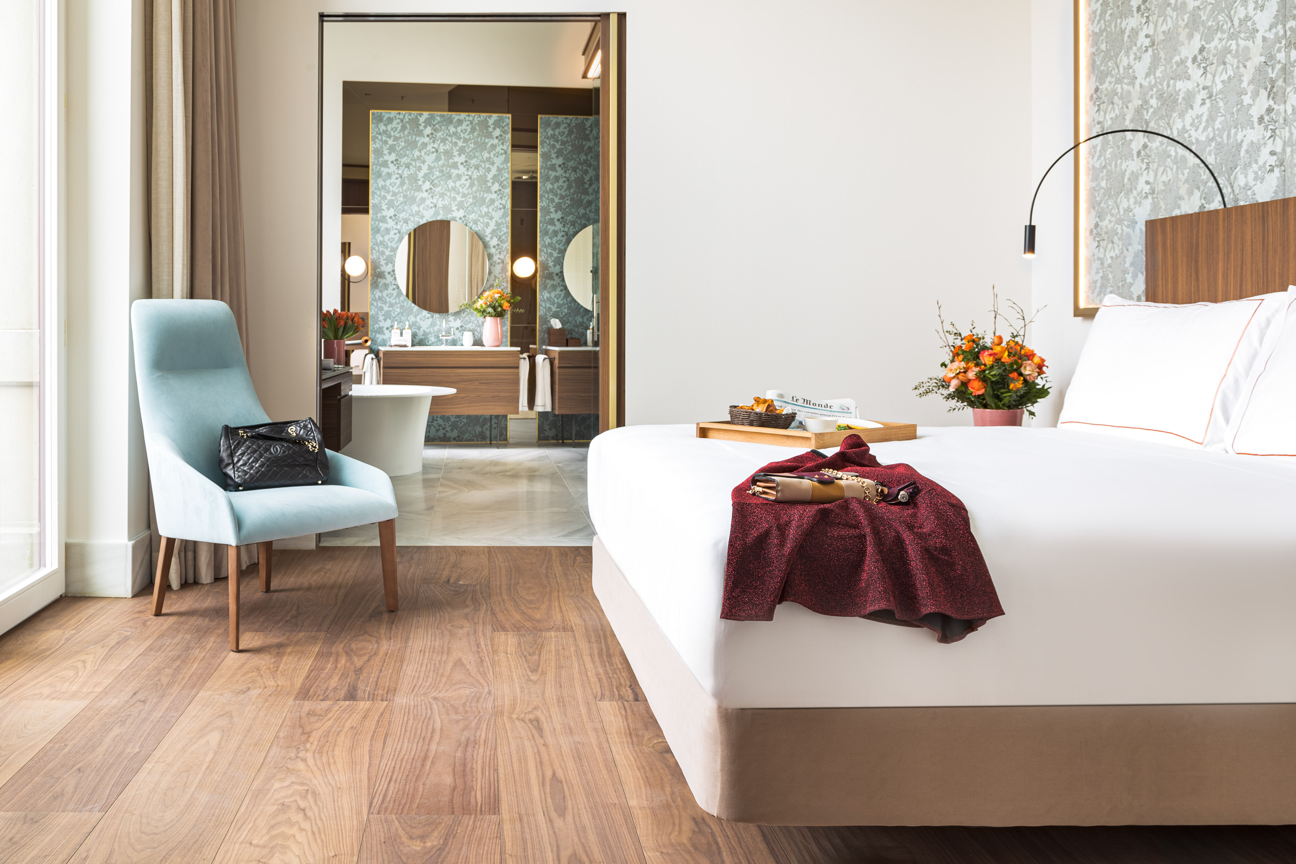 Luxury hotel bedroom with king bed, accent chair, wooden floor, open bathroom.
