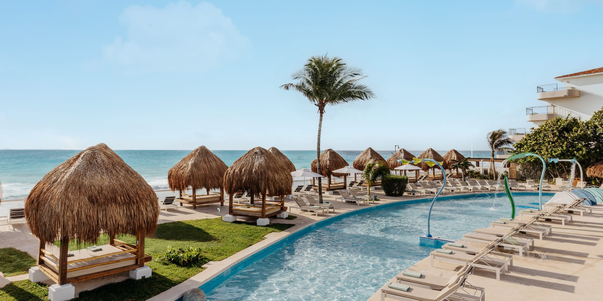 Tropical resort pool with ocean view, thatched cabanas, and lounge chairs.
