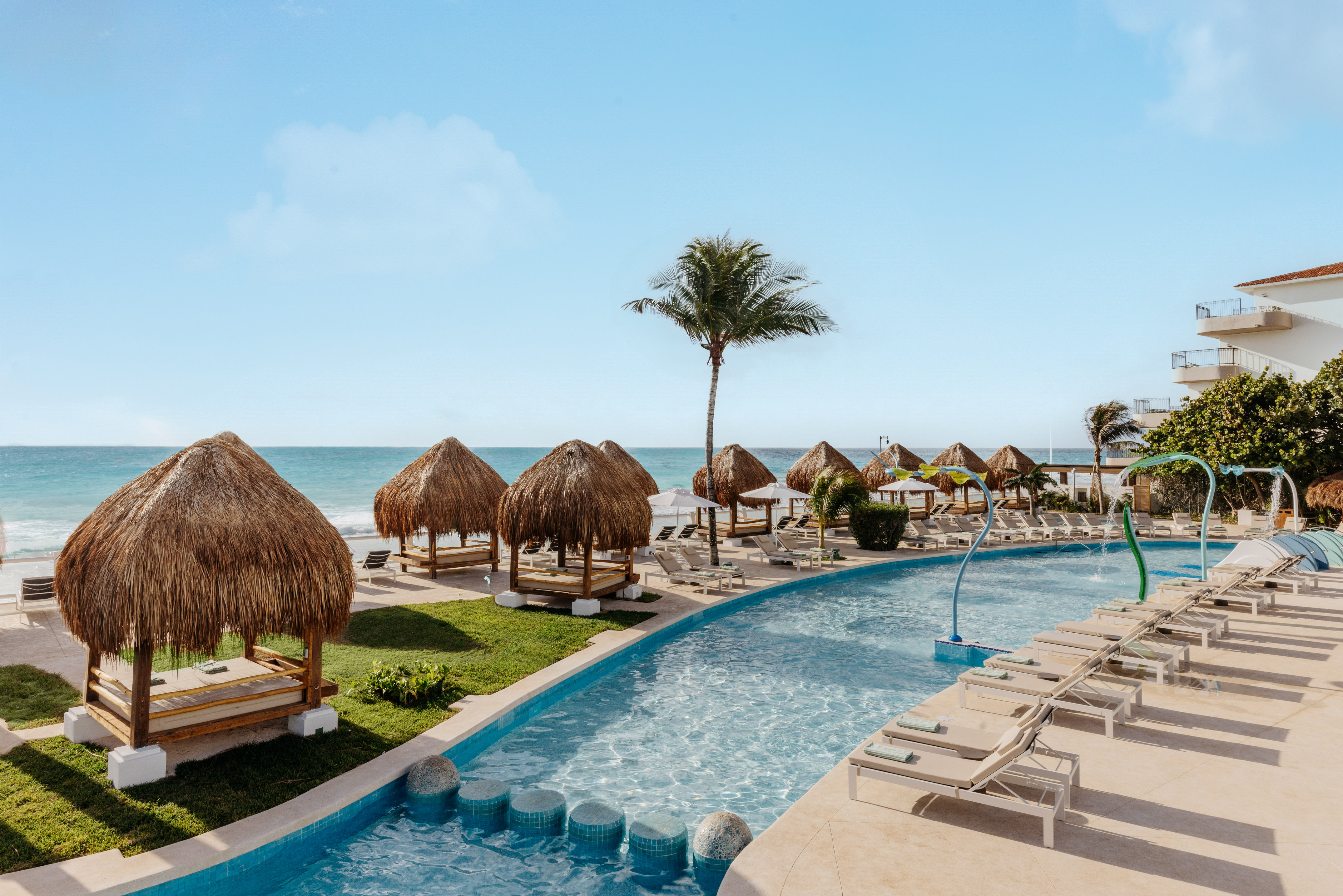 Tropical resort pool with ocean view, thatched cabanas, and lounge chairs.
