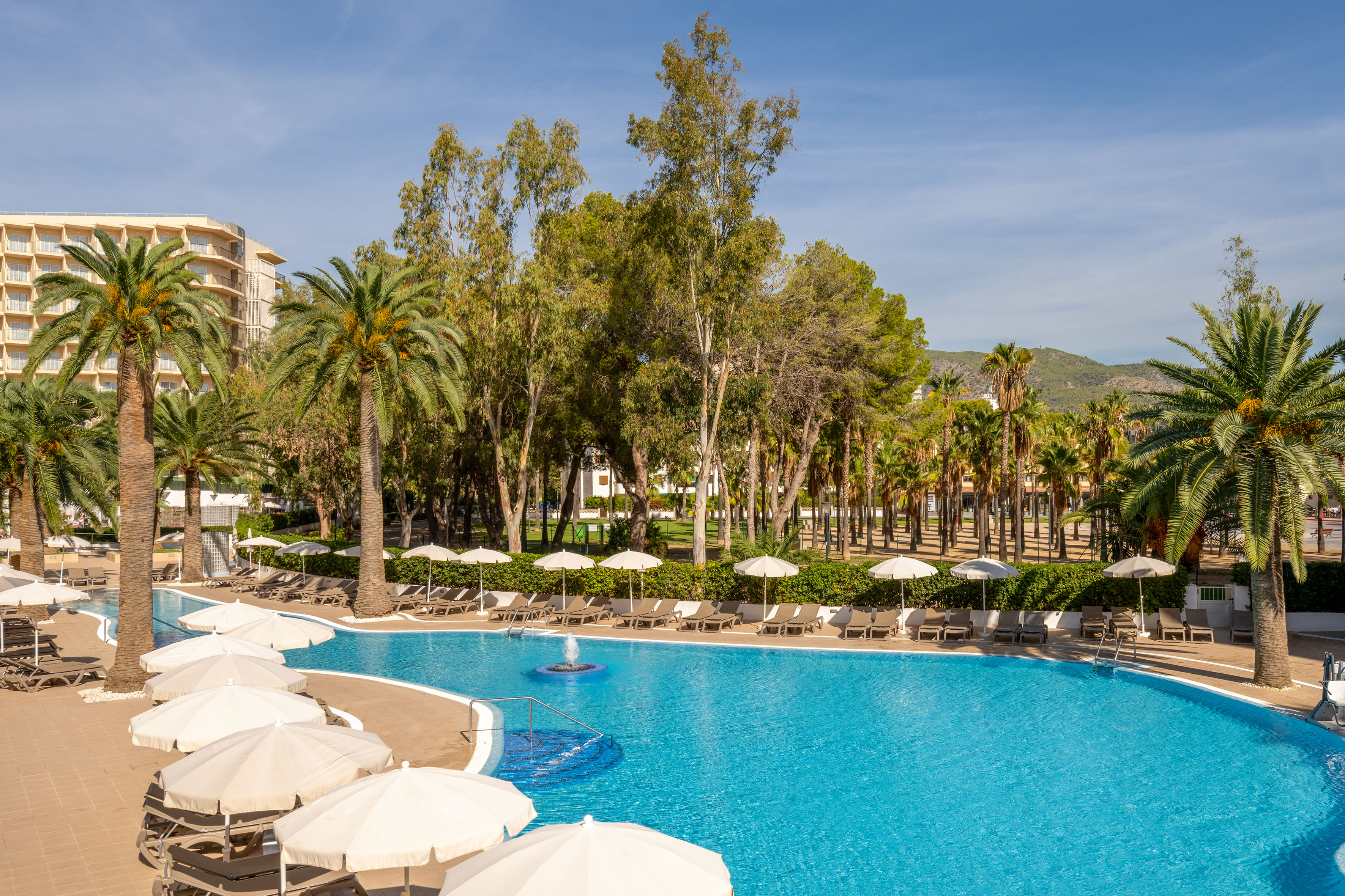 Hotel resort pool with sun loungers, umbrellas, palm trees, blue sky.