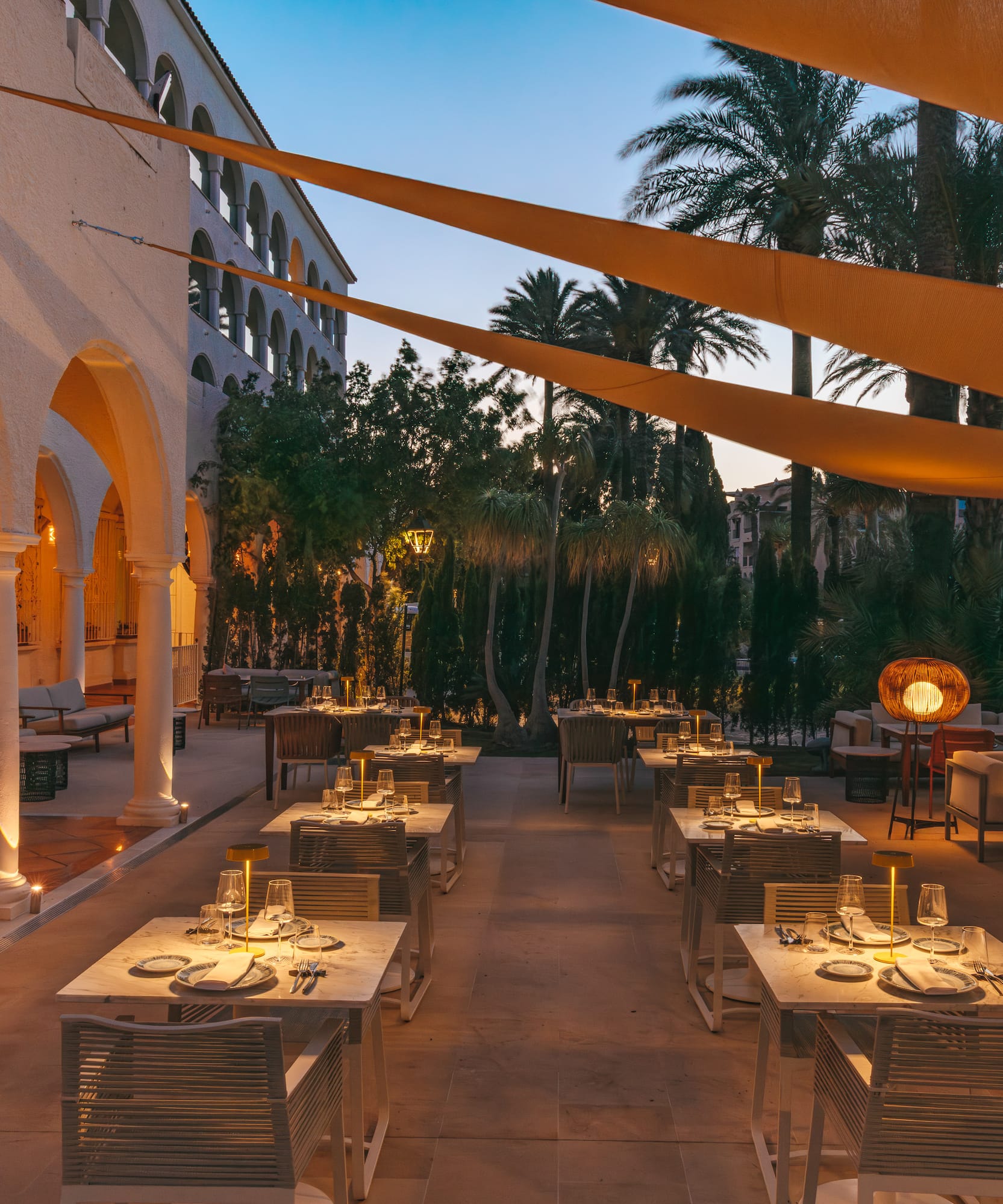 Elegant al fresco dining terrace with evening lighting, set tables, and palm trees.