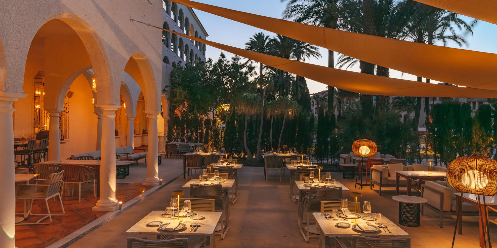 Elegant al fresco dining terrace with evening lighting, set tables, and palm trees.
