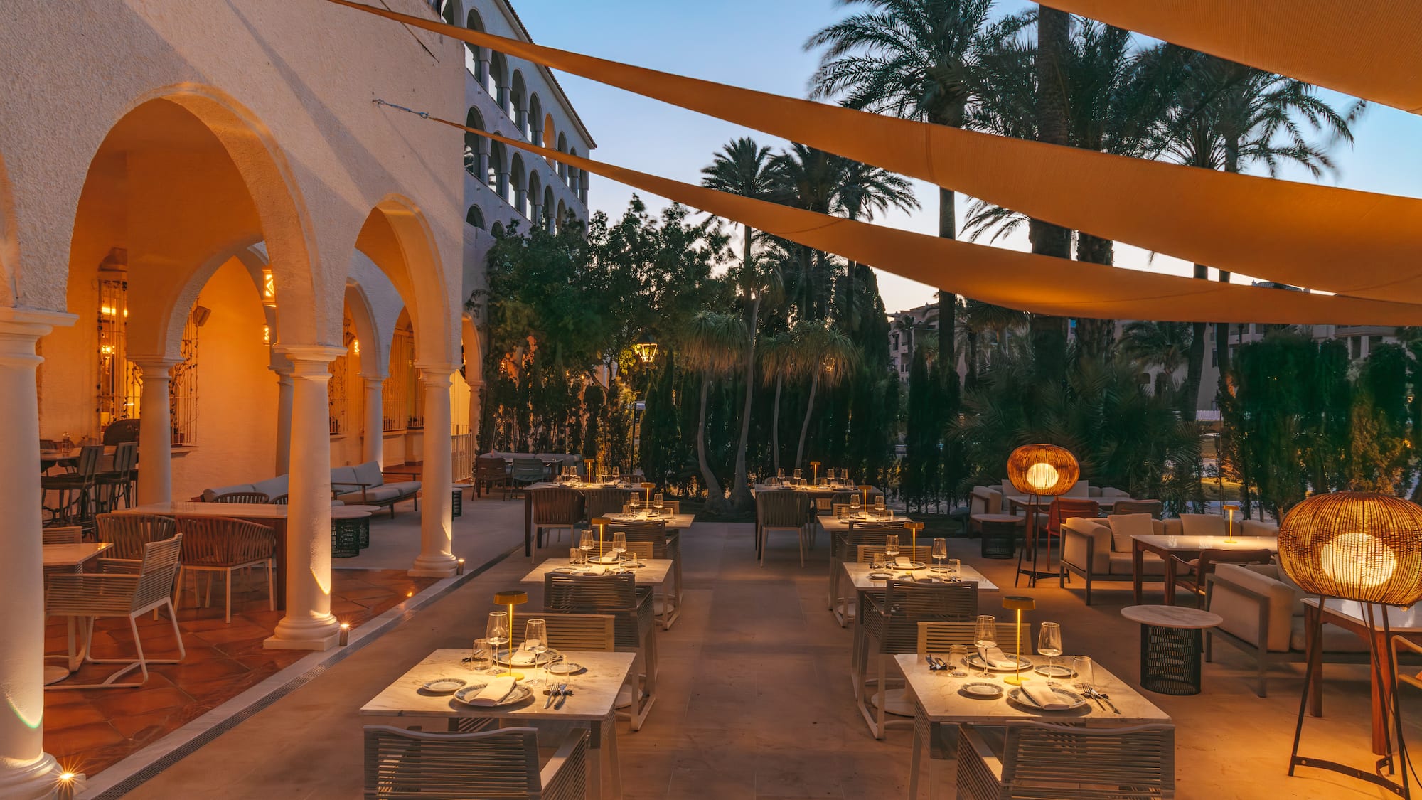Outdoor restaurant terrace at twilight, set tables, palm trees, warm lighting.