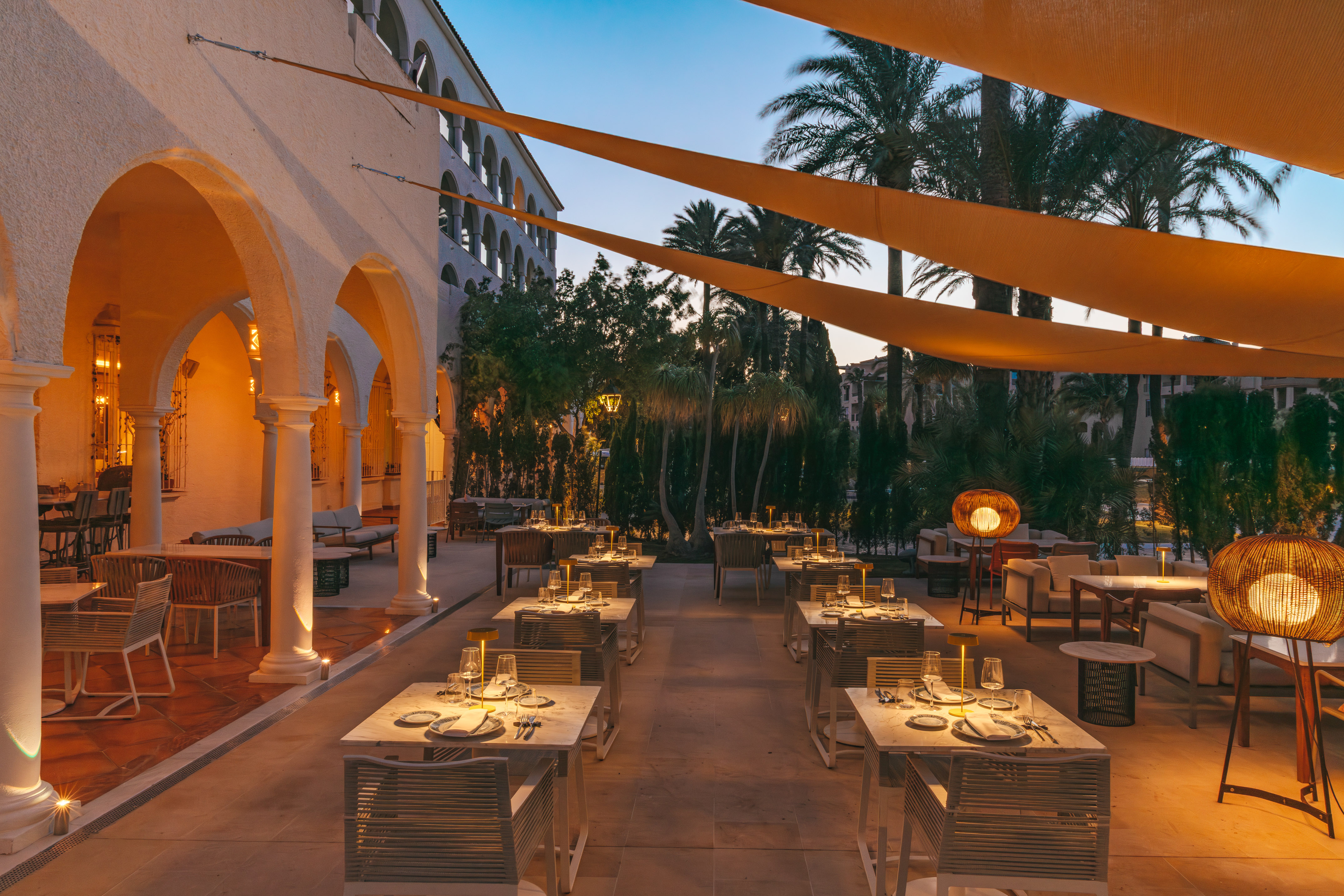 Elegant al fresco dining terrace with evening lighting, set tables, and palm trees.