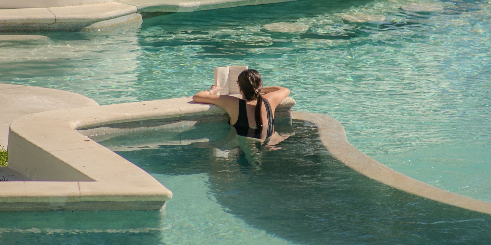 Woman in black swimsuit reading a book in a clear swimming pool.