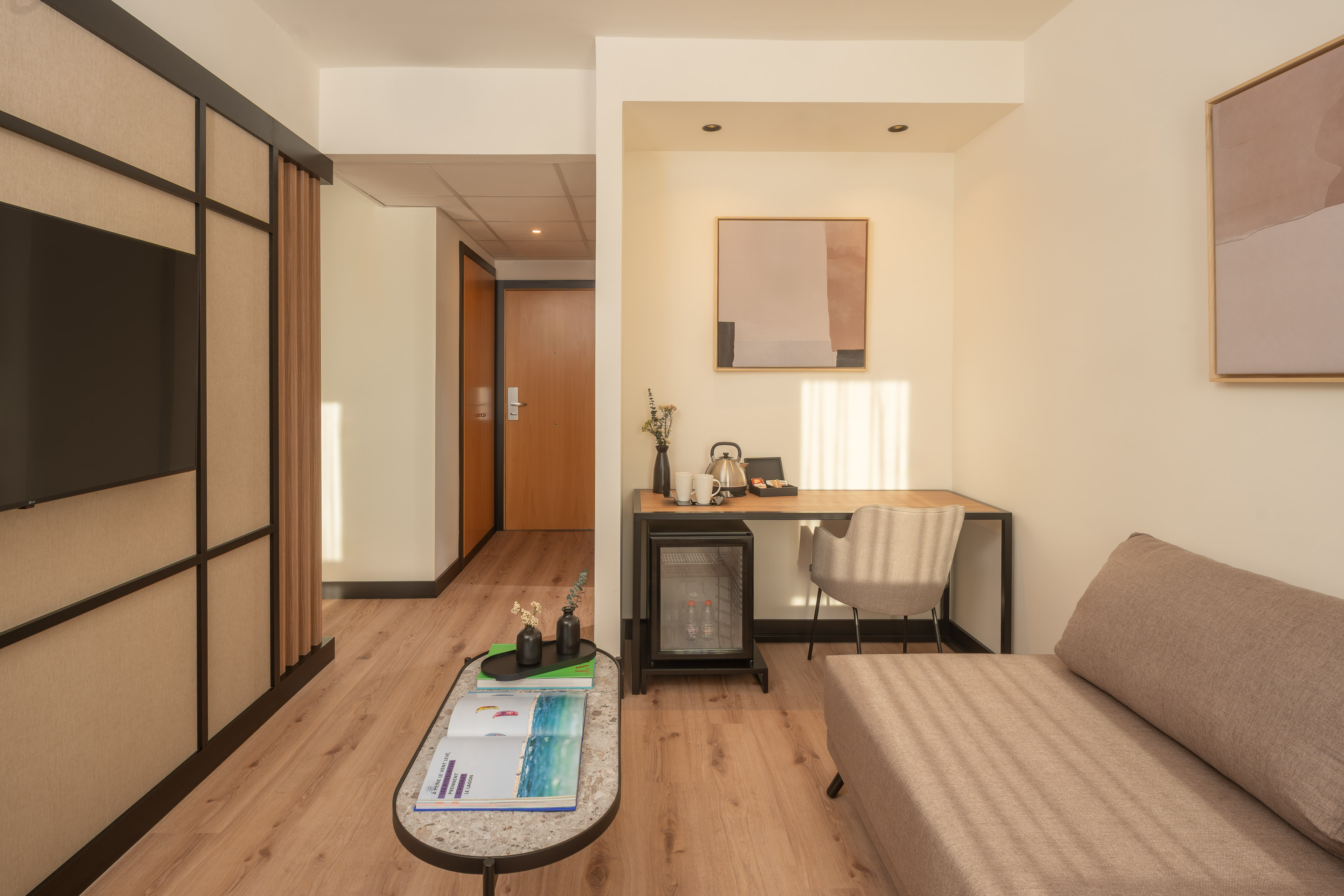 Modern hotel room with sofa, desk, minibar, TV, and wood floor.
