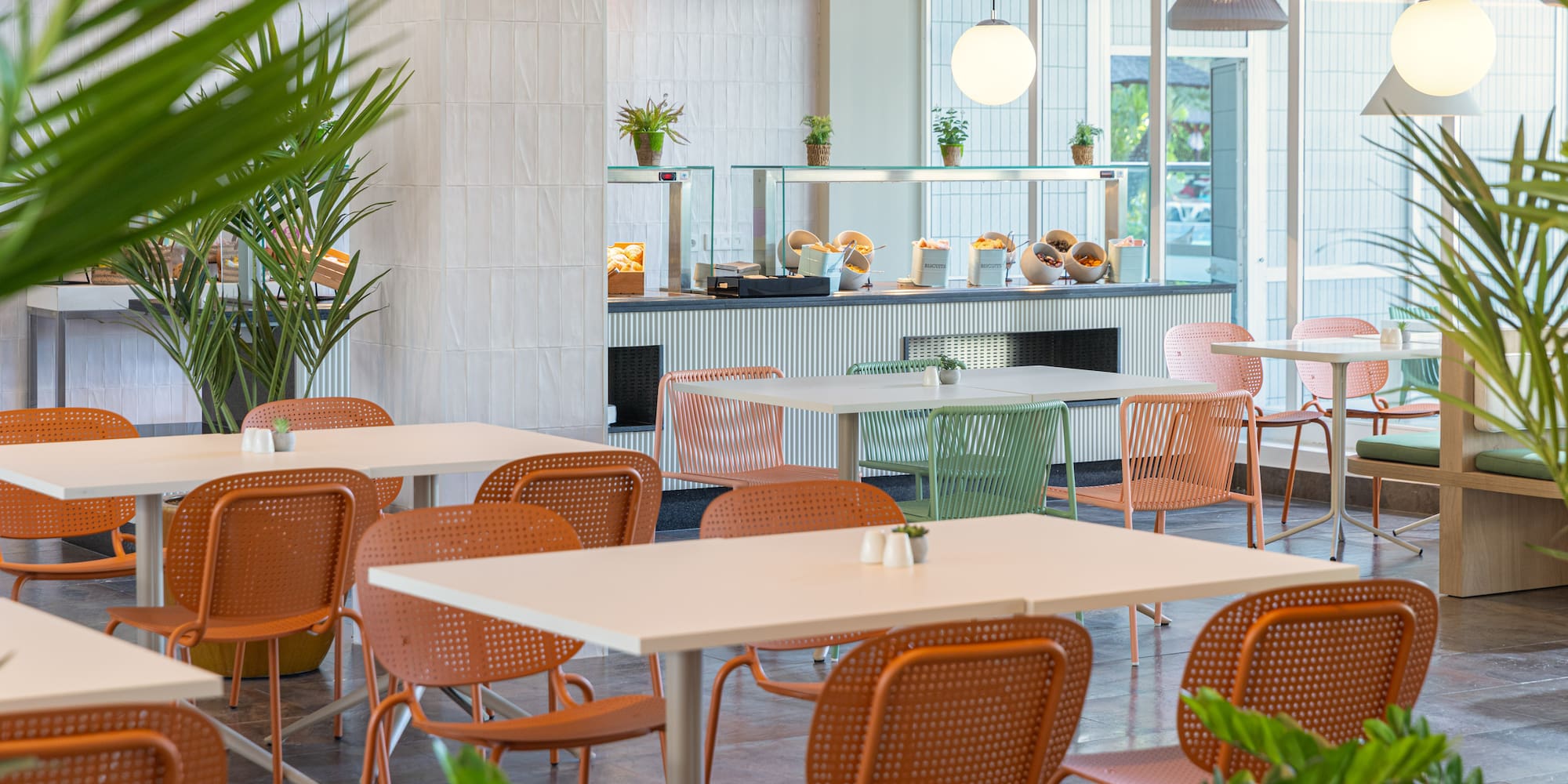 Modern hotel breakfast buffet with colorful chairs, natural light, and plants.