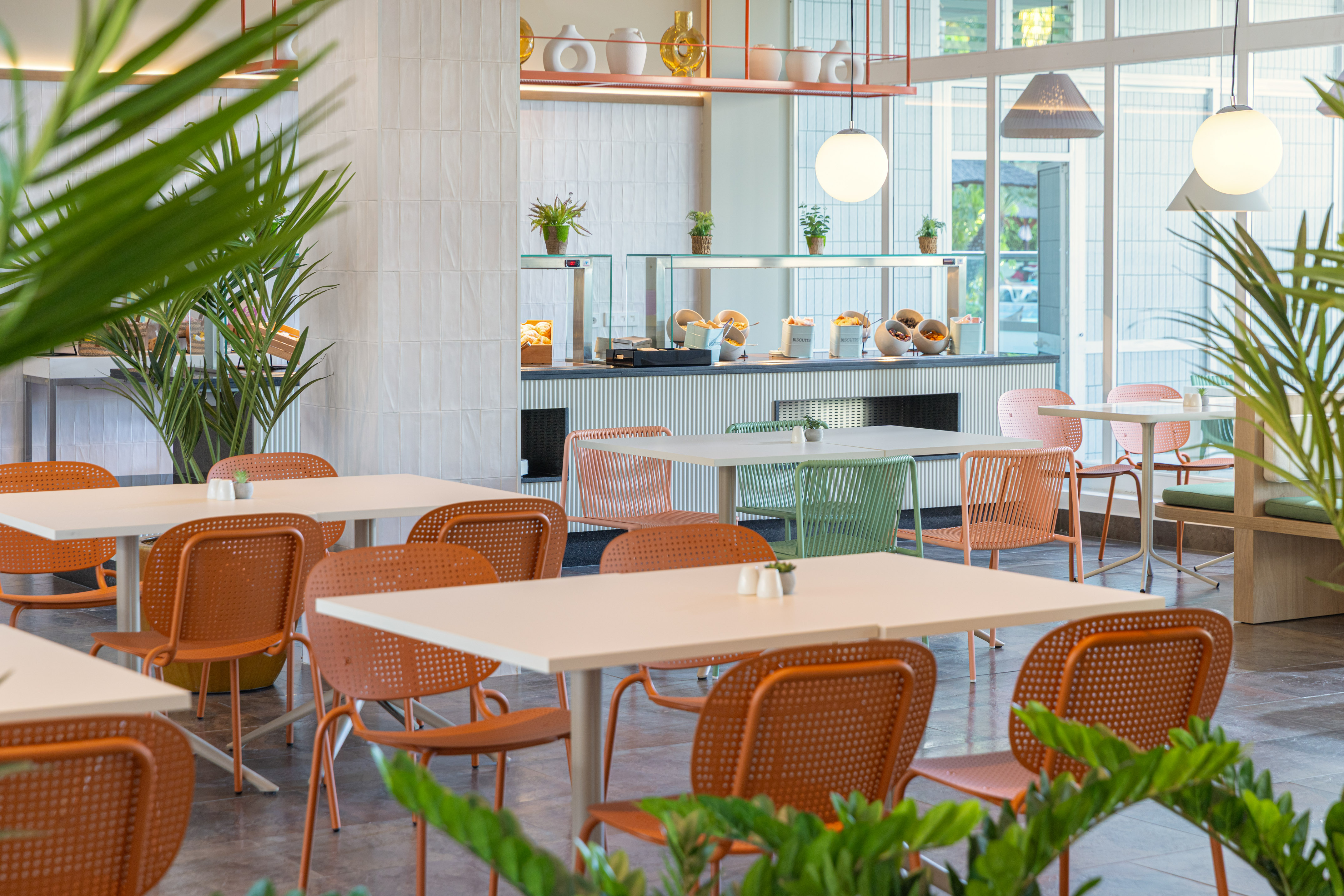 Modern hotel breakfast buffet with colorful chairs, natural light, and plants.