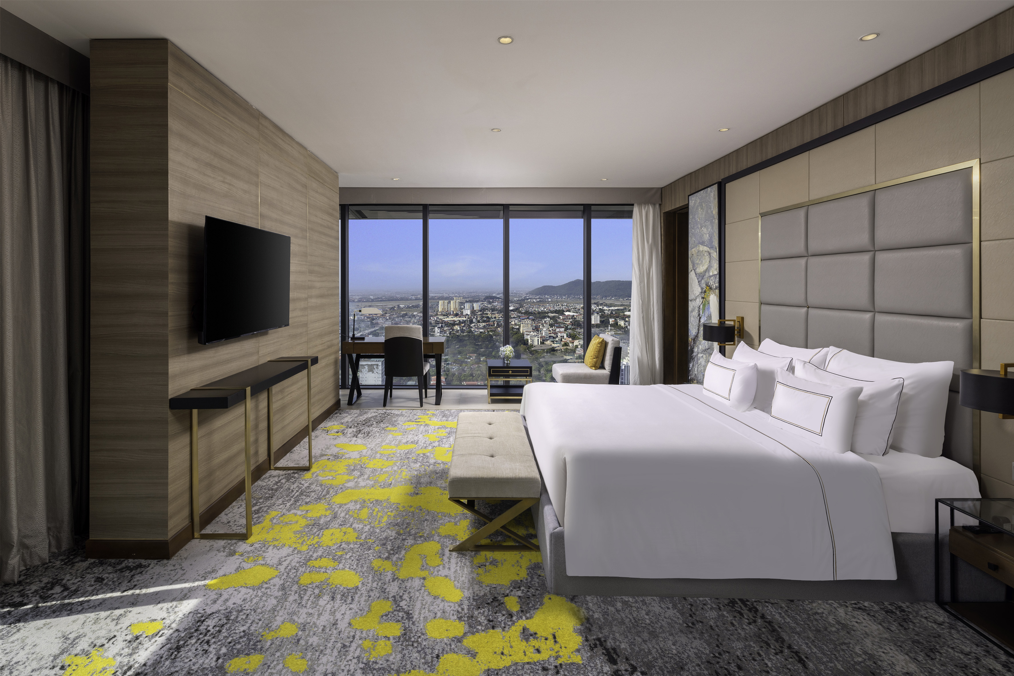 Modern hotel bedroom, king bed, panoramic city view, desk, yellow-patterned carpet.