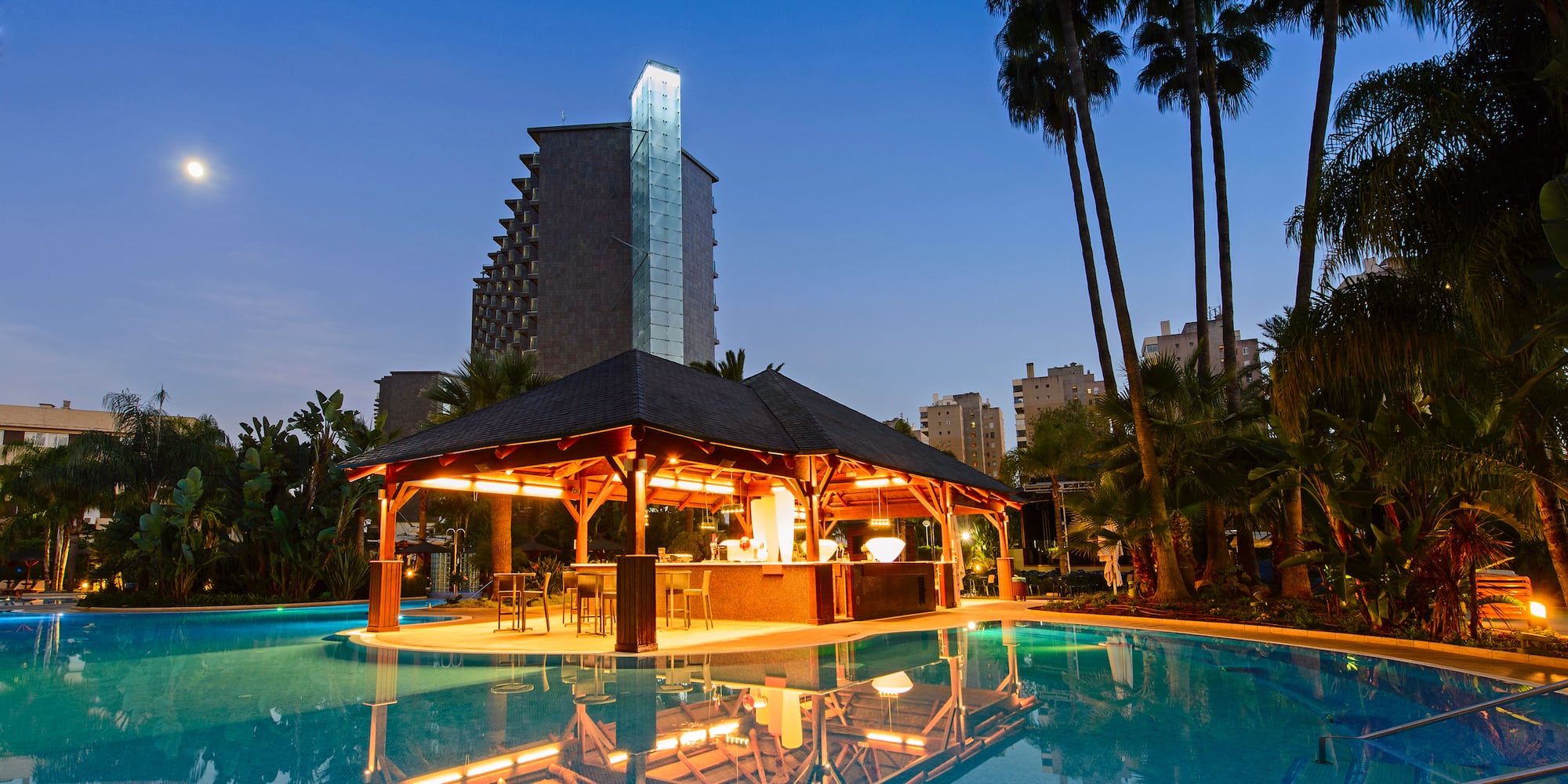 Illuminated resort pool bar under a moonlit sky with palm trees.