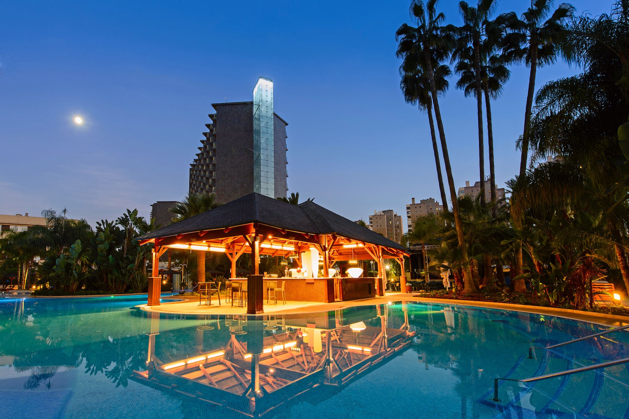 Illuminated resort pool bar under a moonlit sky with palm trees.