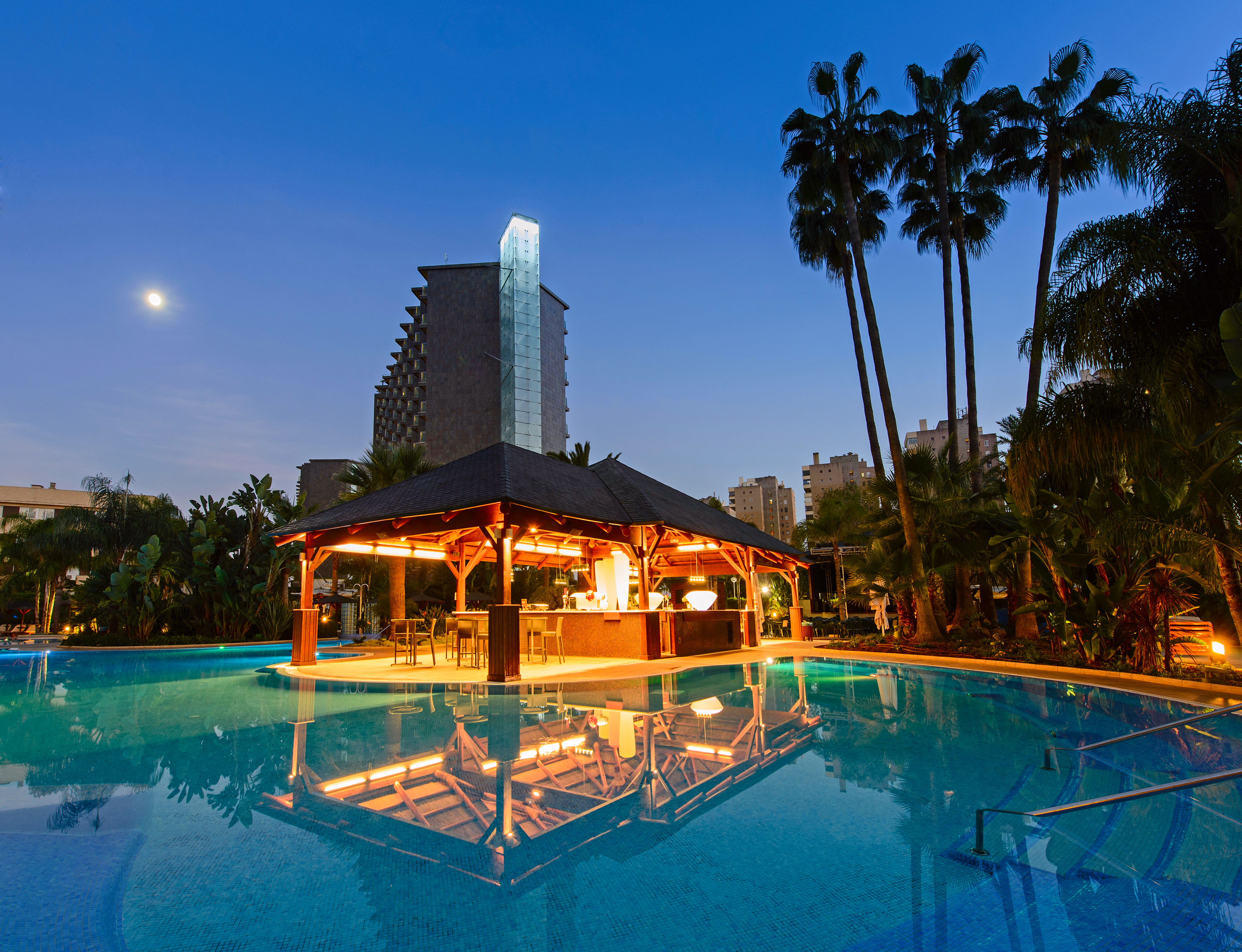 Illuminated resort pool bar under a moonlit sky with palm trees.