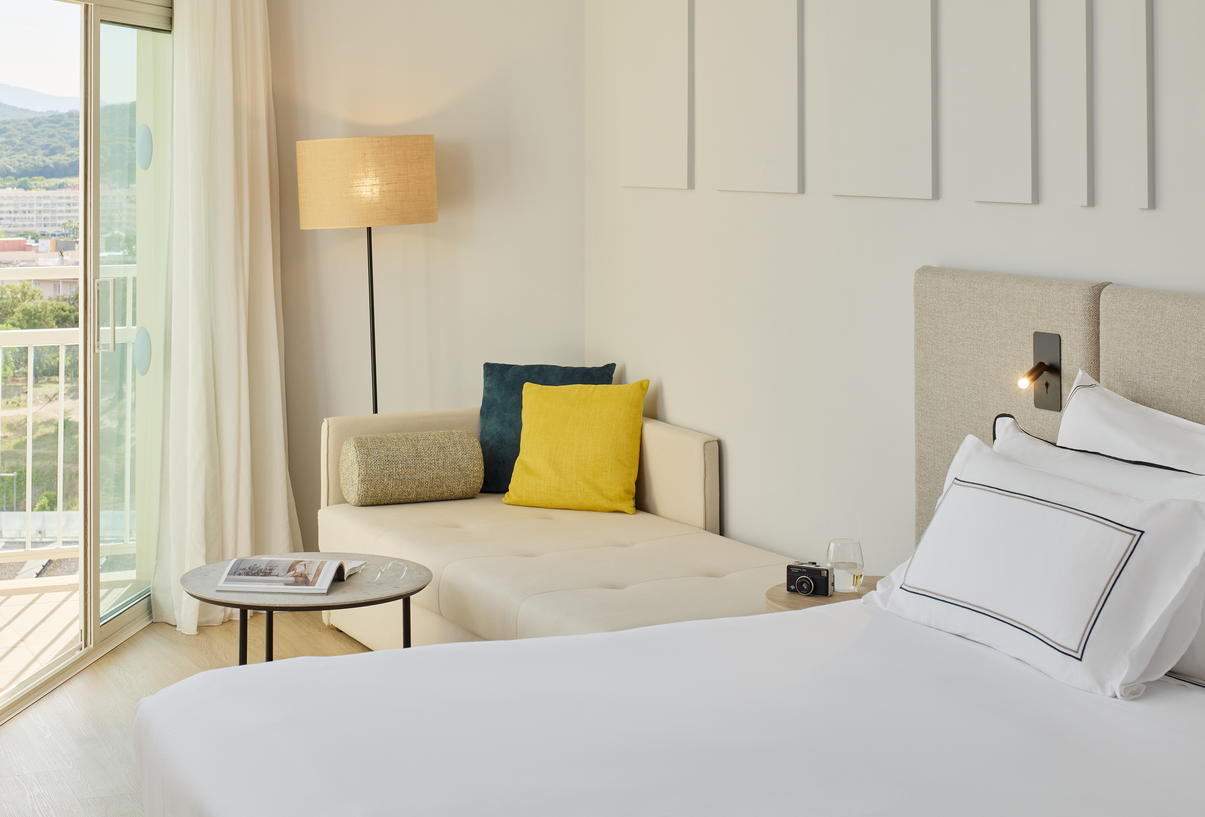 Bright hotel room with bed, chaise lounge, colorful pillows, and balcony.