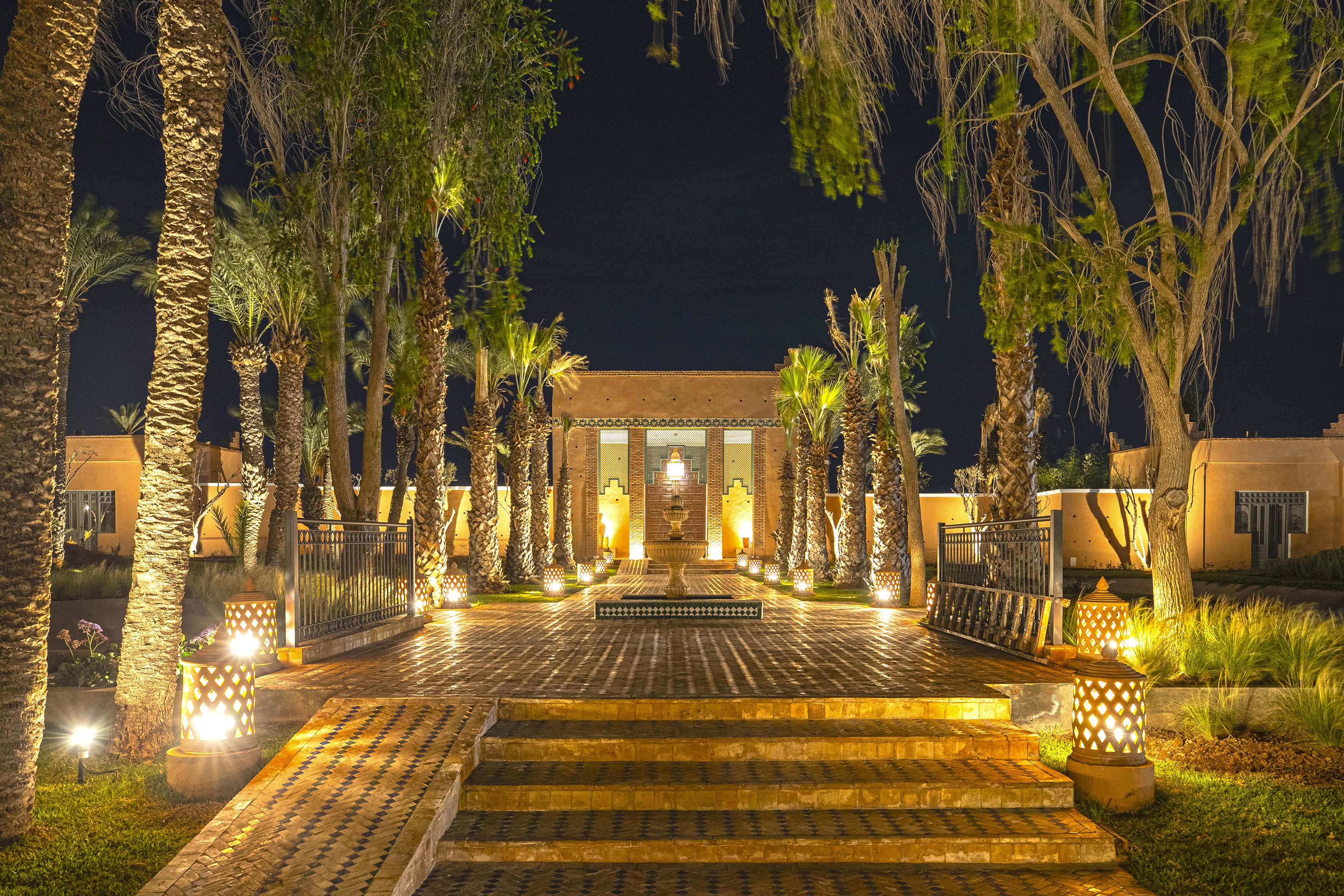 Nightlit Moroccan courtyard with palm trees, ornate fountain, and tiled steps.