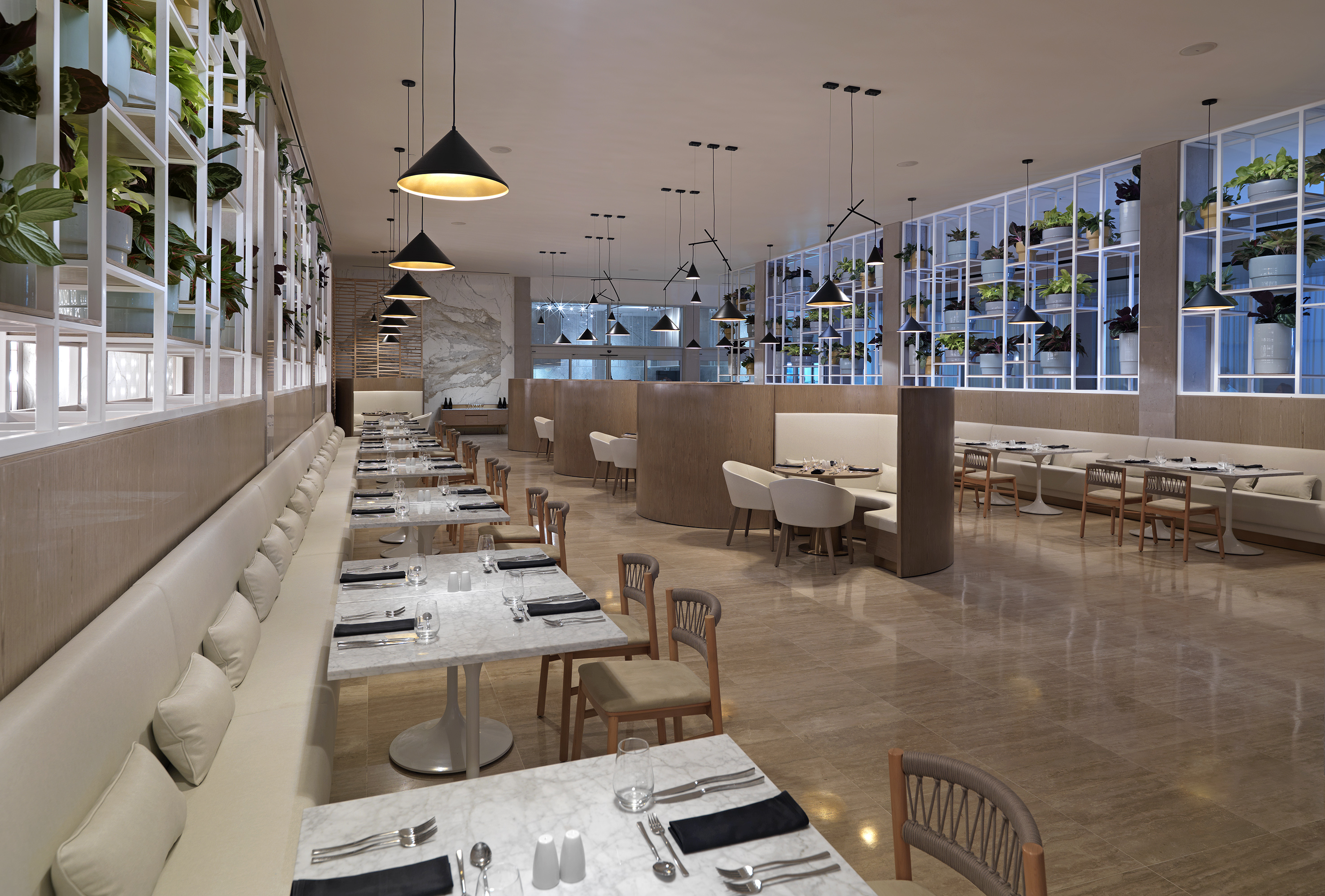 Modern restaurant interior with white marble tables, green plants, and pendant lights.