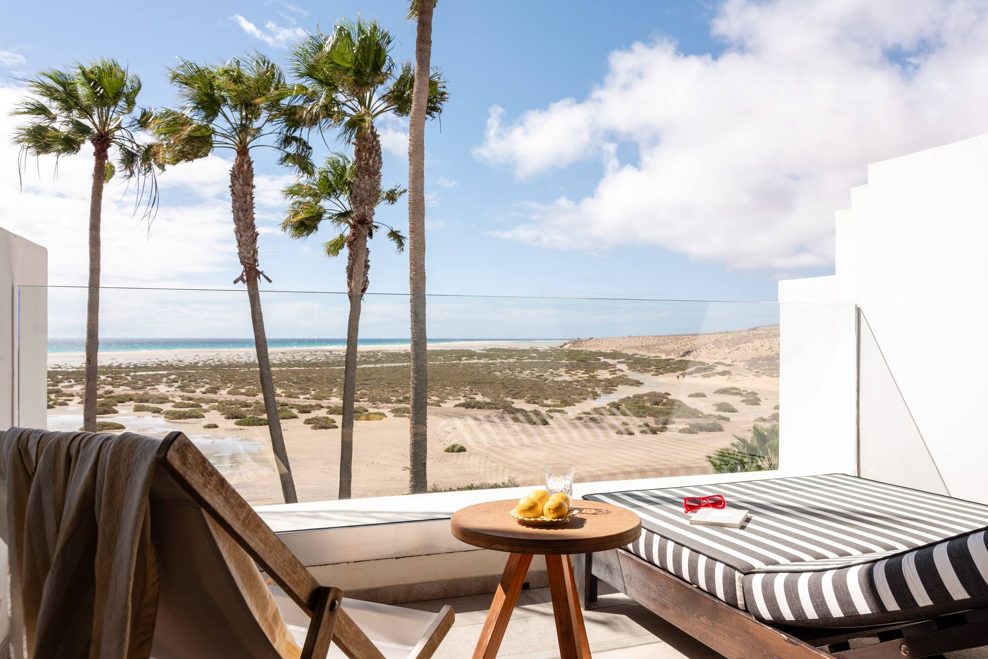 Sunny balcony overlooking tropical beach with palm trees and lounge furniture.
