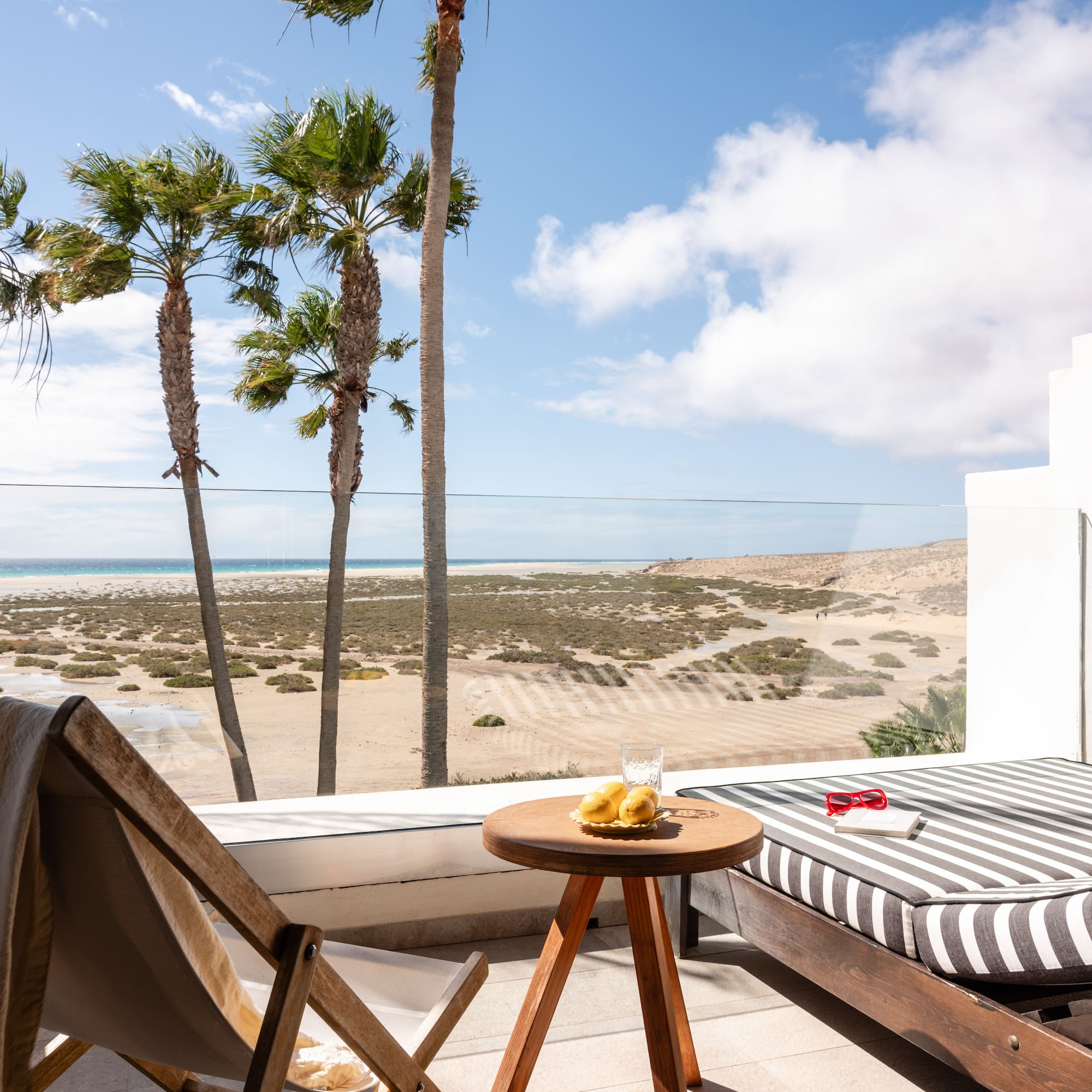 Sunny balcony overlooking tropical beach with palm trees and lounge furniture.