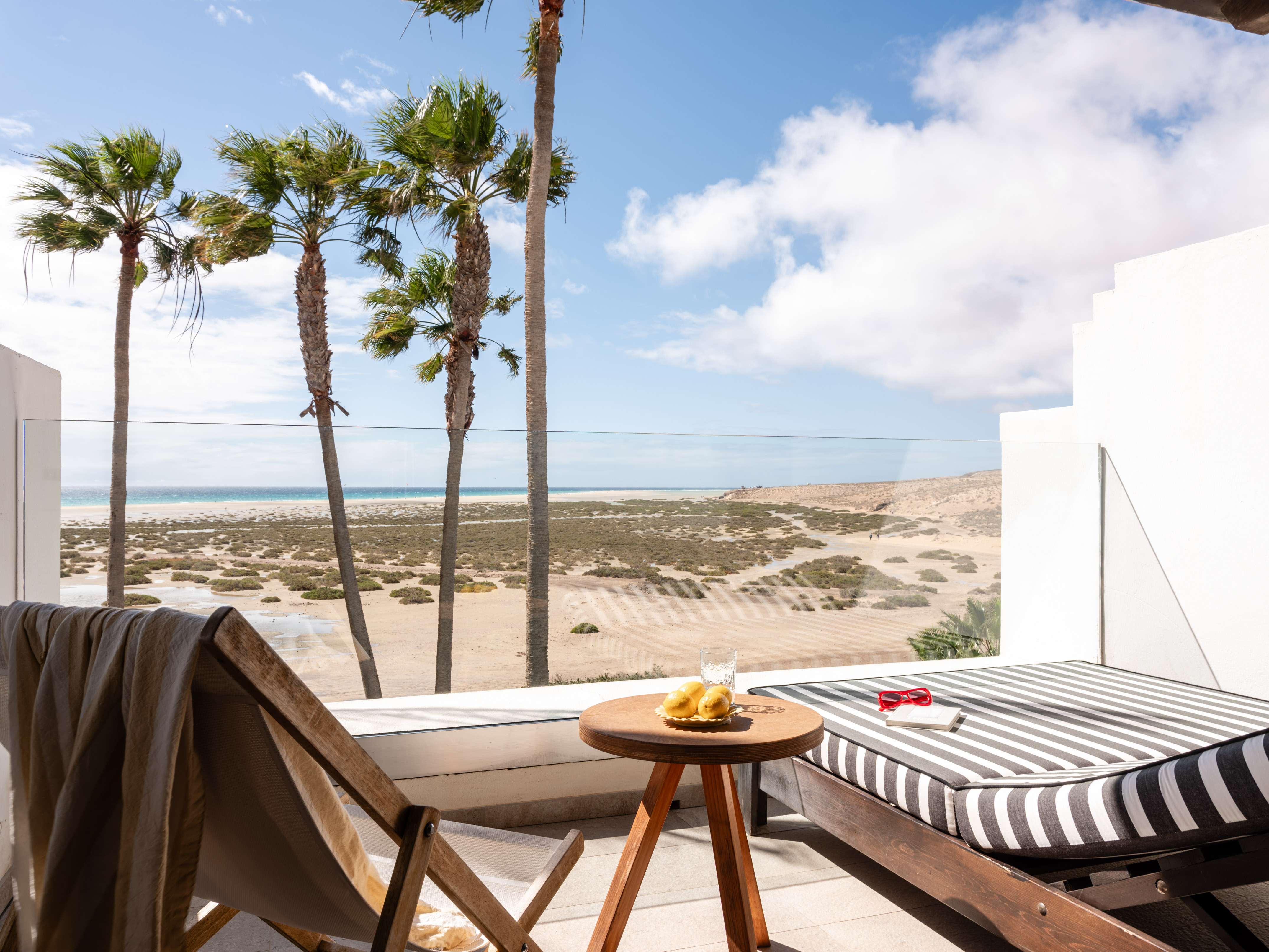 Sunny balcony overlooking tropical beach with palm trees and lounge furniture.