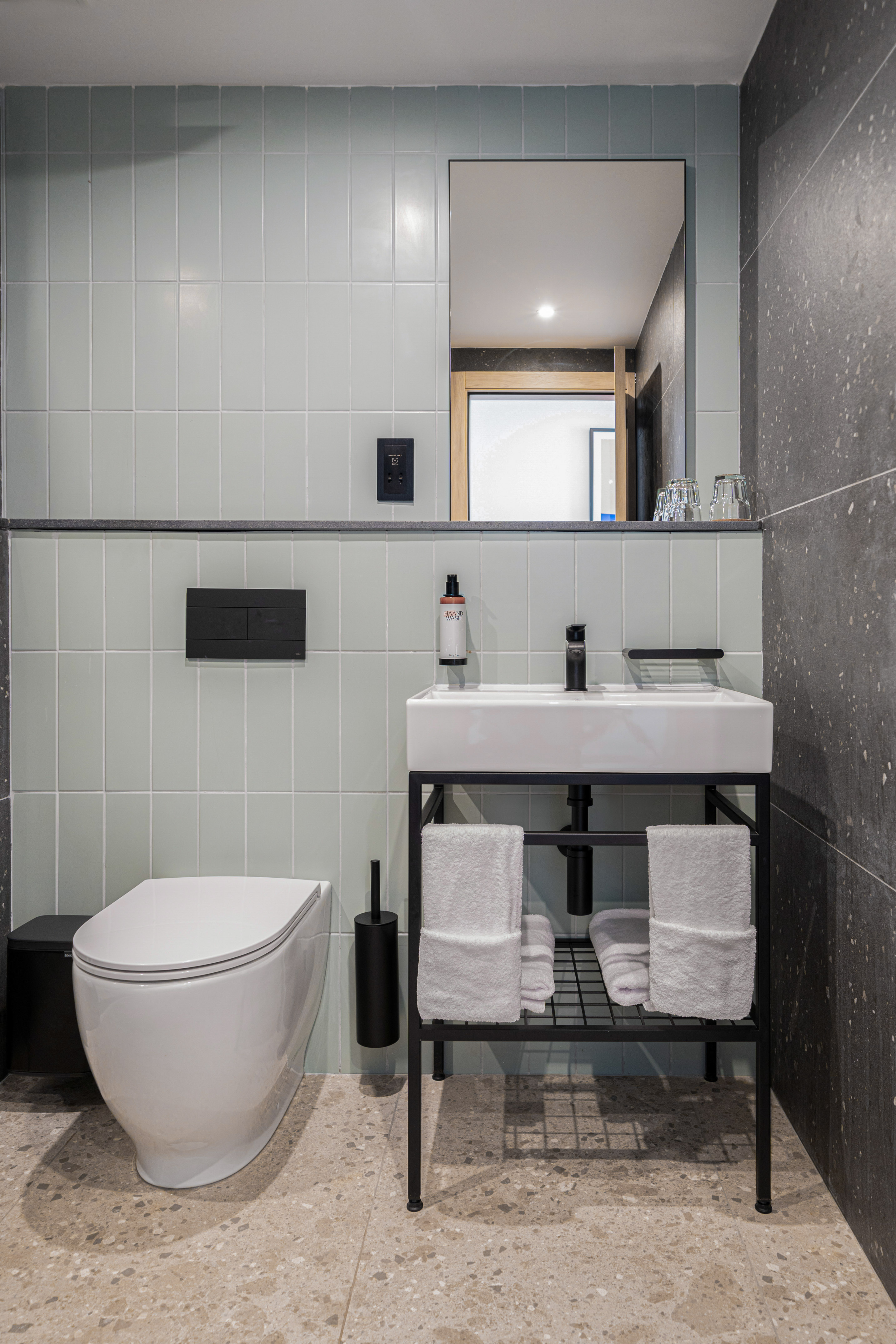 Modern hotel bathroom with light blue tiles, white toilet, sink, and terrazzo floor.