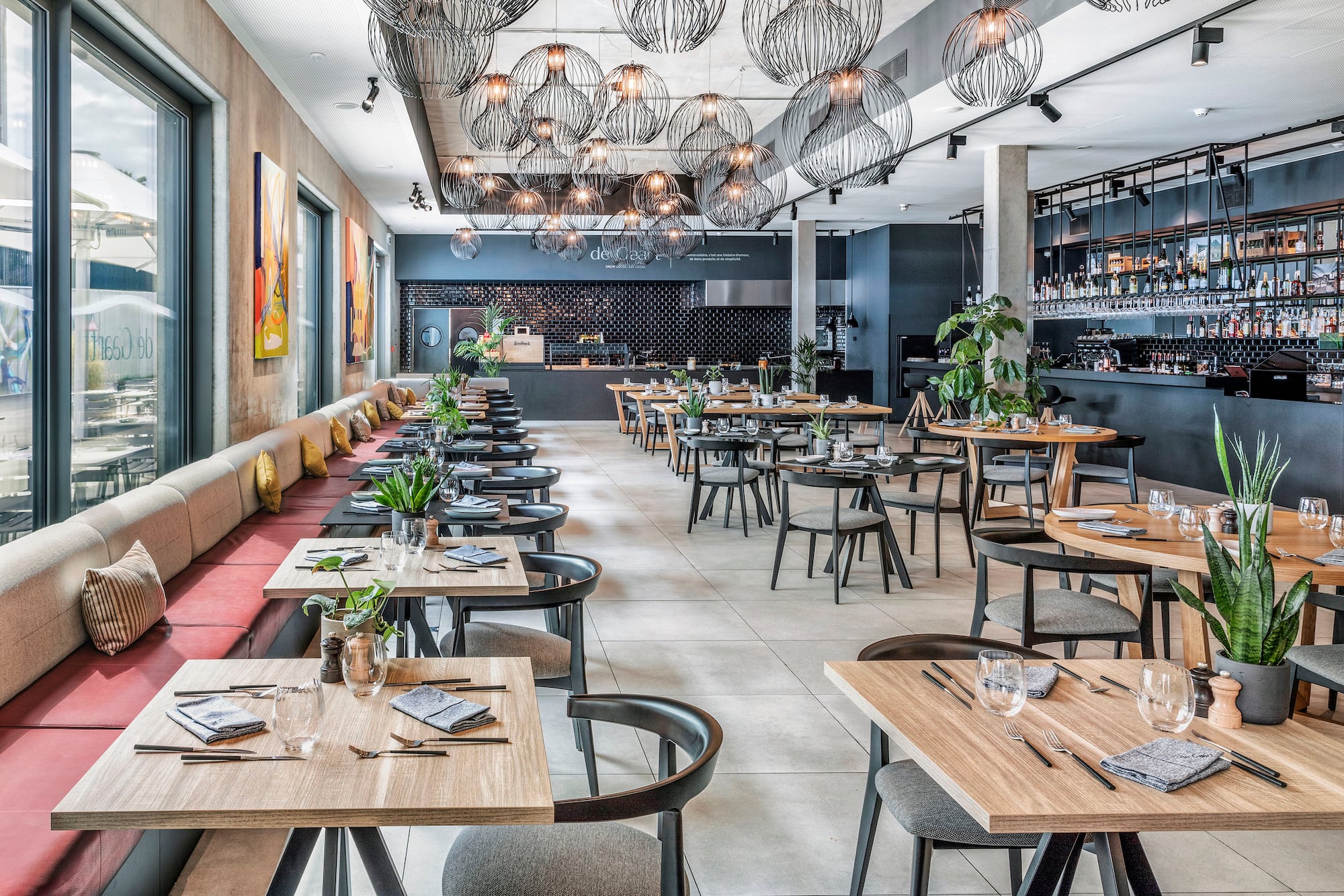 Modern restaurant dining room with tables, banquette seating, bar, and pendant lights.