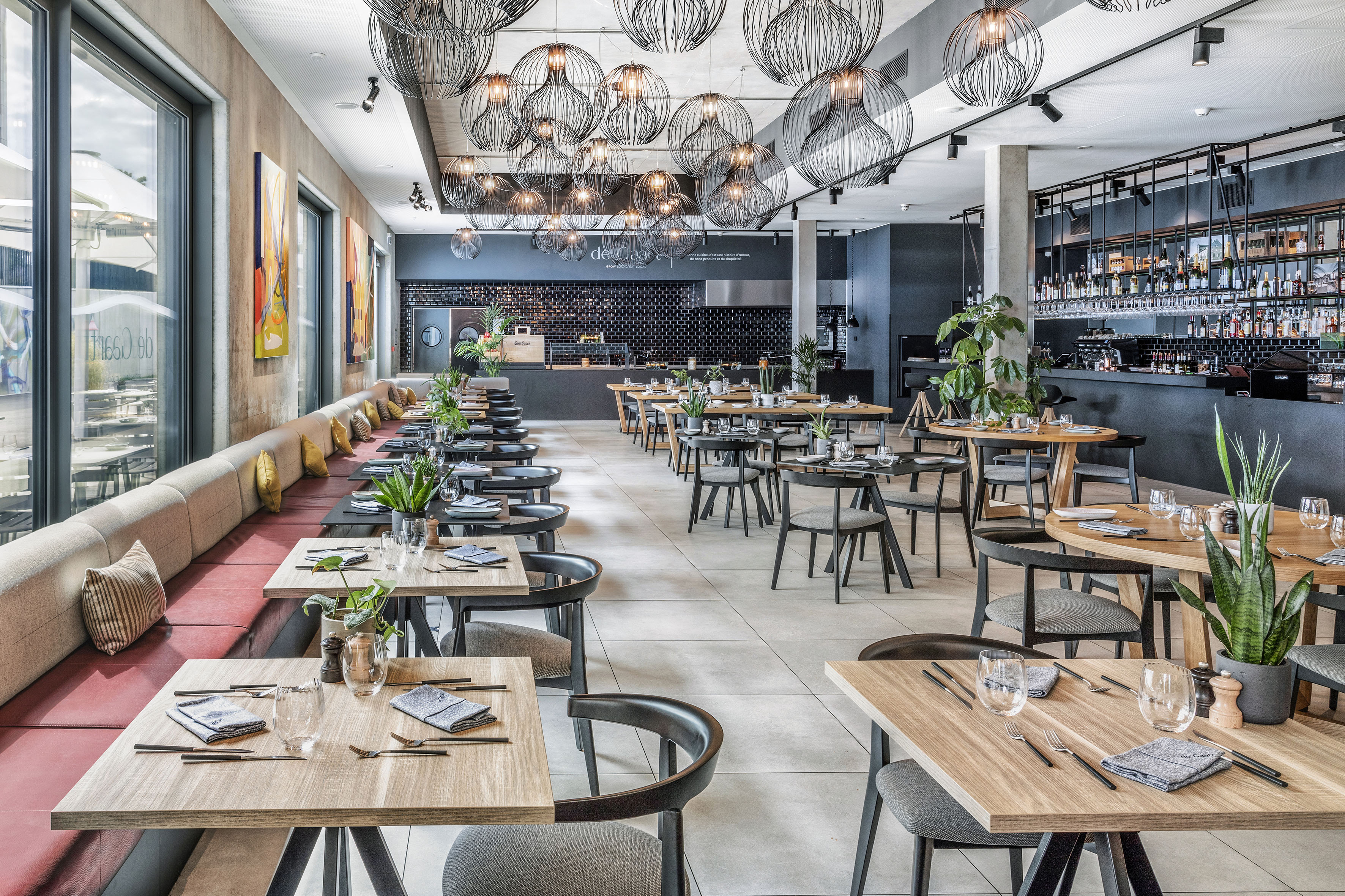 Modern restaurant dining room with tables, banquette seating, bar, and pendant lights.
