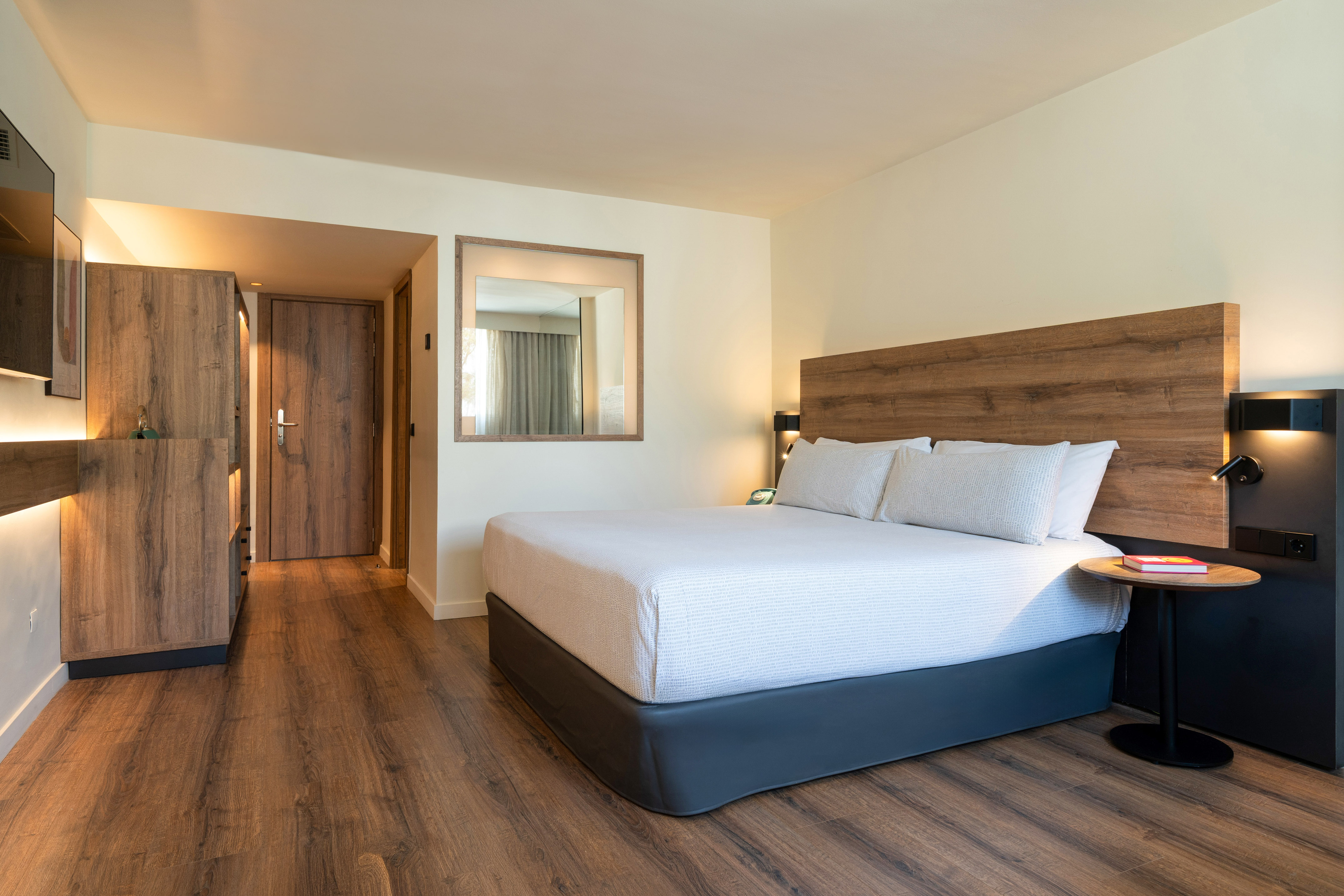 Modern hotel room with bed, wooden headboard, side table, and wood floor.
