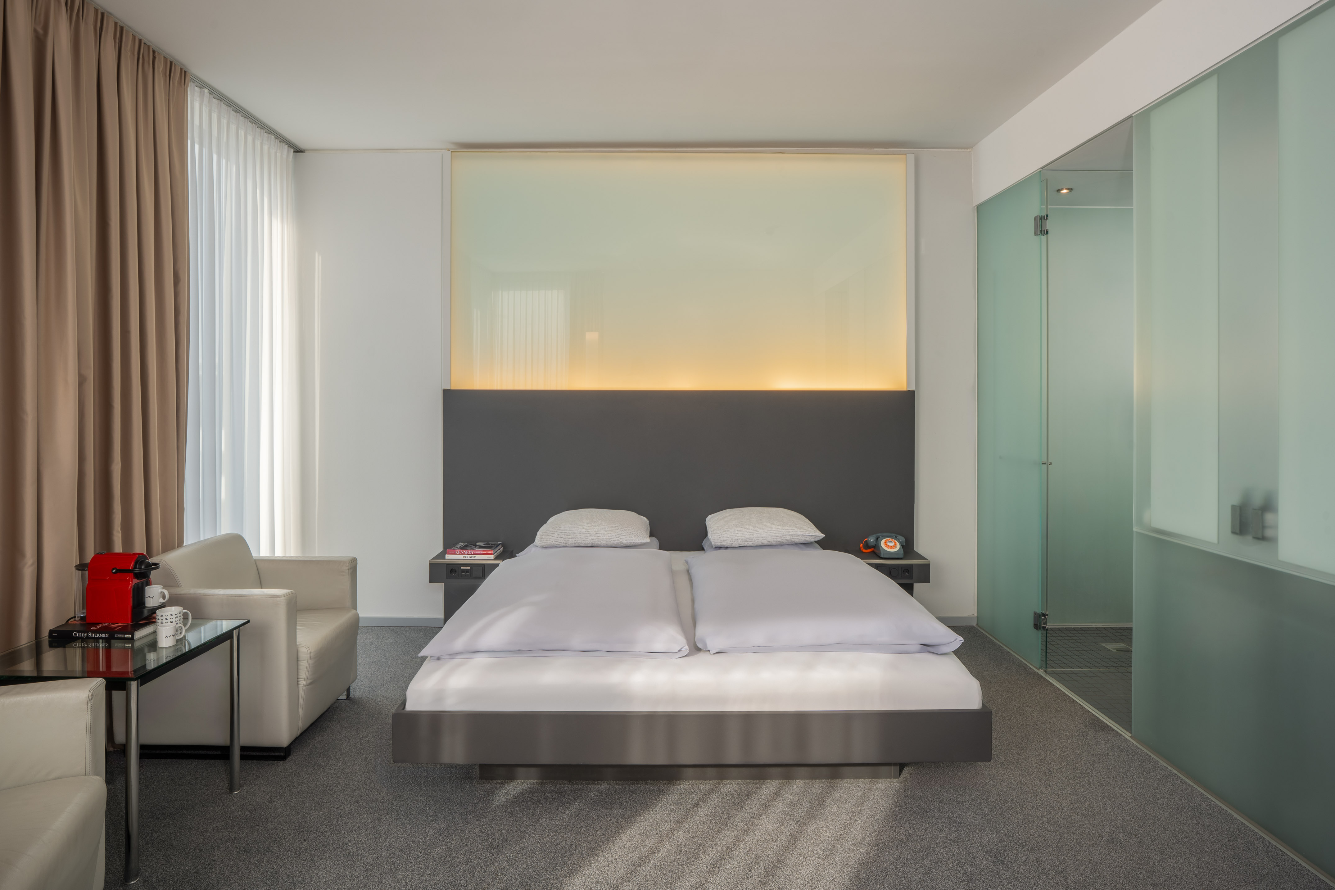 Modern hotel bedroom with king bed, backlit headboard, and frosted glass shower.