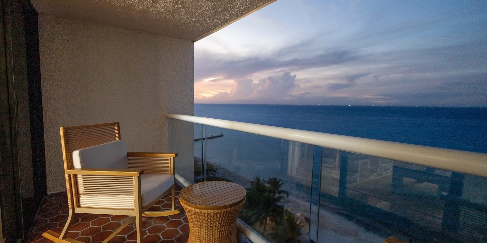 Oceanfront balcony with rocking chair, wicker table, sunset ocean view.