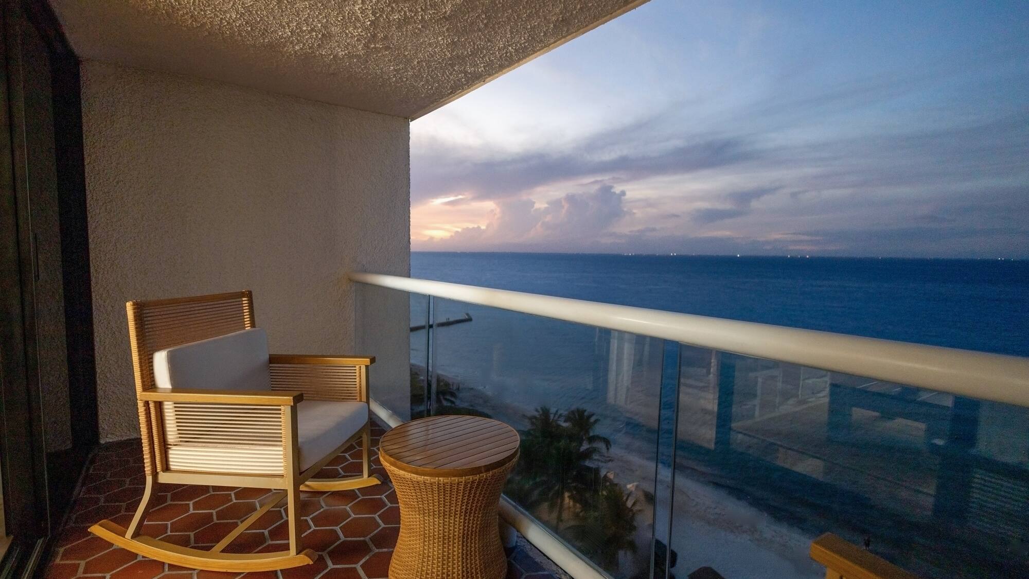 Oceanfront hotel balcony with rocking chair, table, and sunset view.